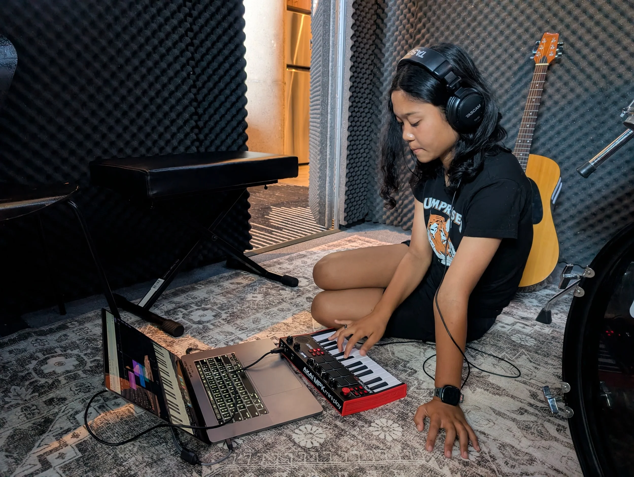 A girl sitting on the floor in a music recording studio, wearing headphones and using a small keyboard controller connected to a laptop, with a guitar hanging on the wall behind her.