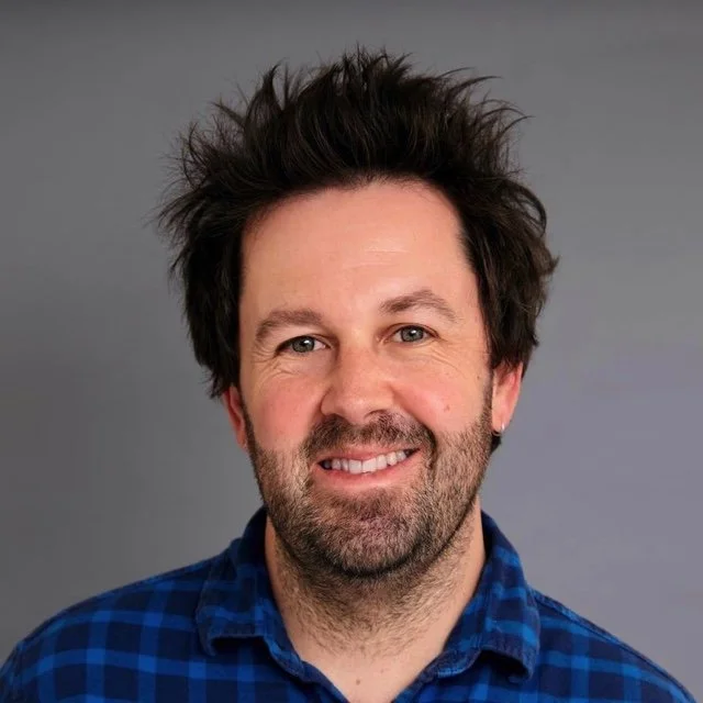 Headshot of a smiling man with curly dark hair, beard, and a small earring, wearing a blue checkered shirt, against a gray background.