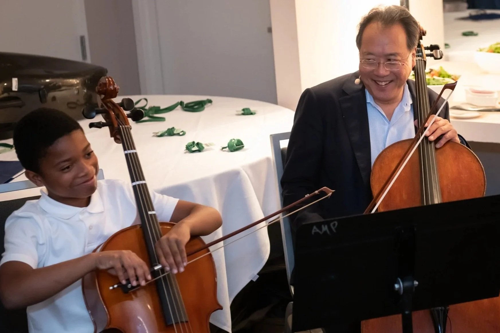 A boy and Yo-Yo Ma are playing cellos at a table, smiling and enjoying music together.