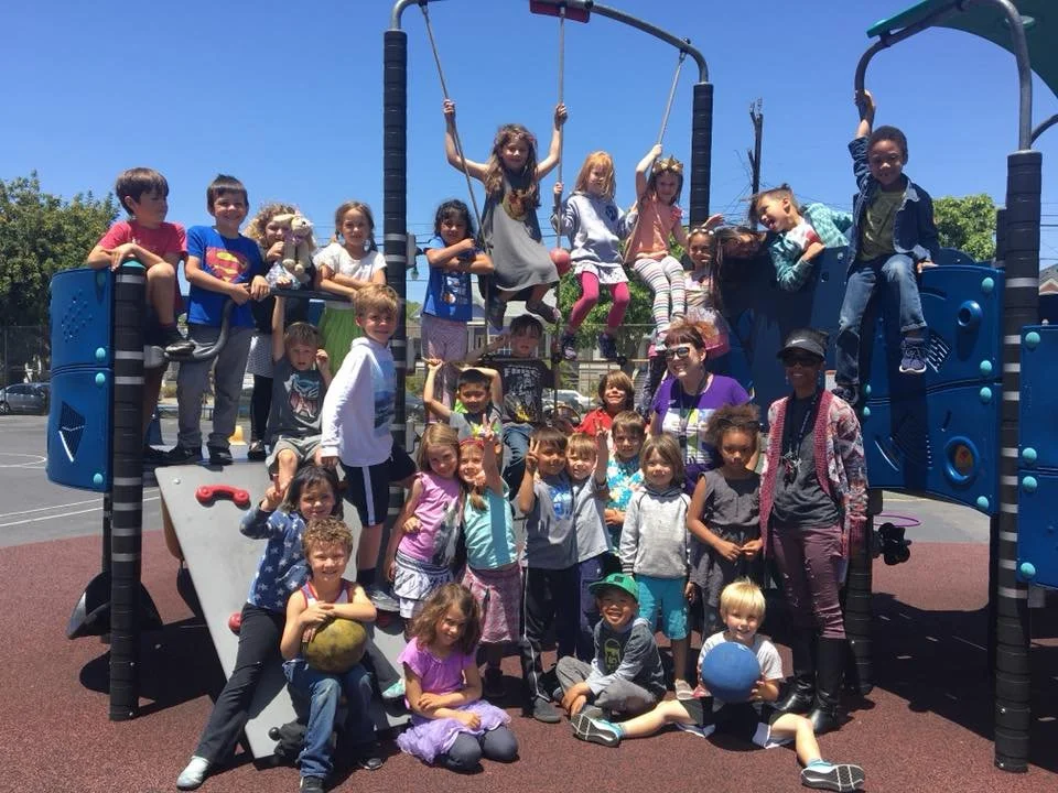 A large group of children and a few adults on a playground structure outdoors, with blue sky and trees in the background.