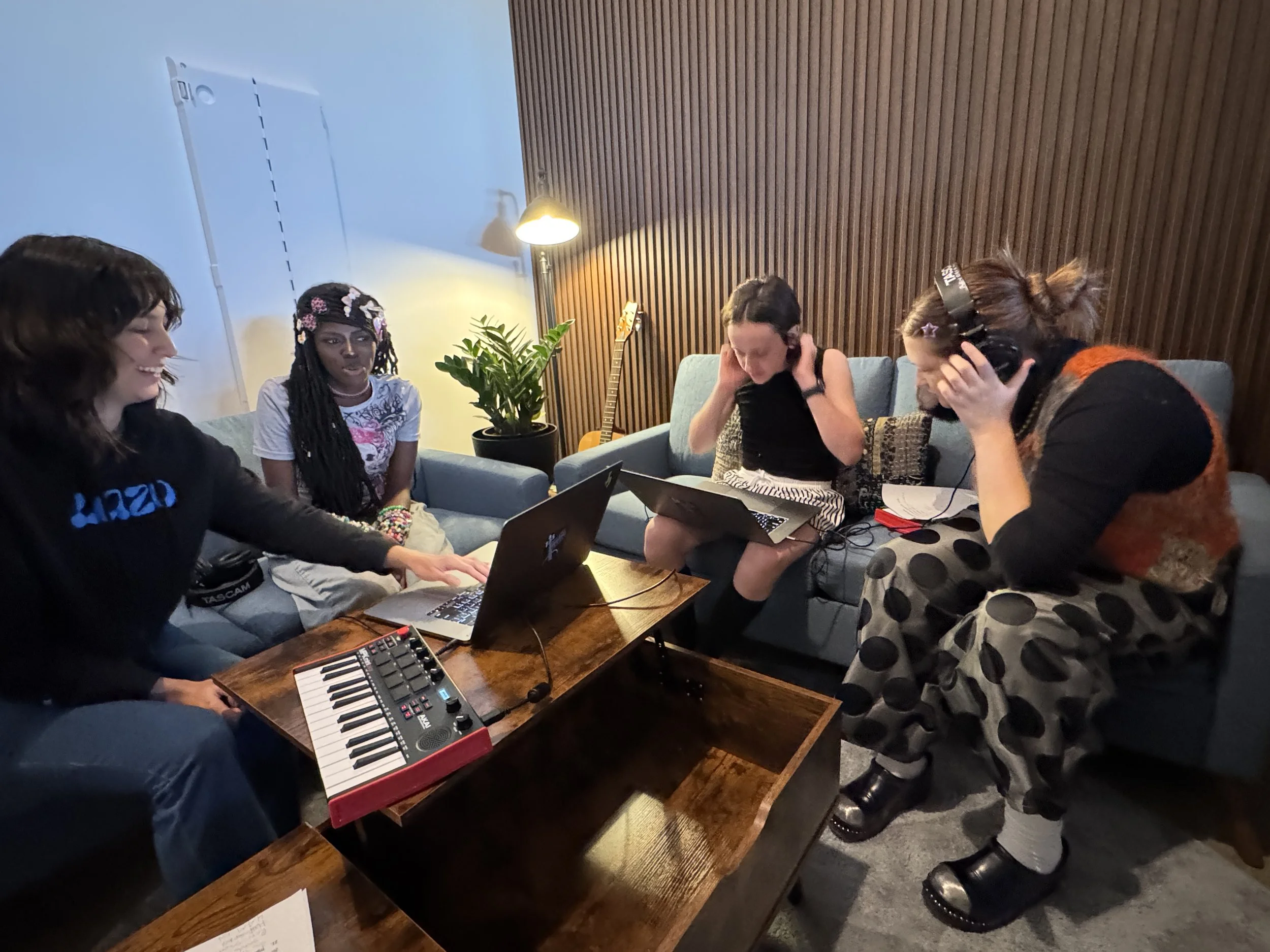 Group of four creators in a music studio, some wearing headphones, with a keyboard, laptop, and recording equipment, engaged in a music session.