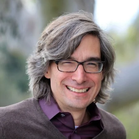 A man with shoulder-length gray hair, glasses, and a big smile, wearing a dark sweater and purple shirt, outdoors in a natural setting.