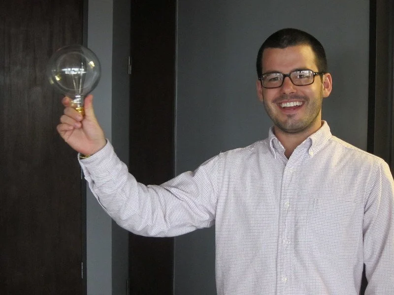 A man with dark hair, glasses, and a trimmed beard is smiling and holding a lightbulb in his right hand, standing indoors next to a dark wall and a door.