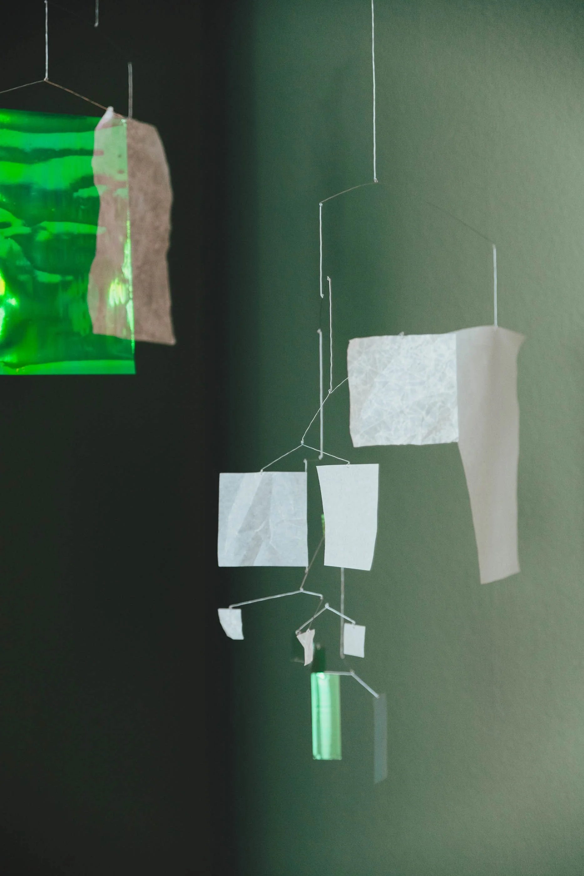 Hängende Kunstinstallation mit weißen und grünen Elementen gegen dunkelgrünen Hintergrund.