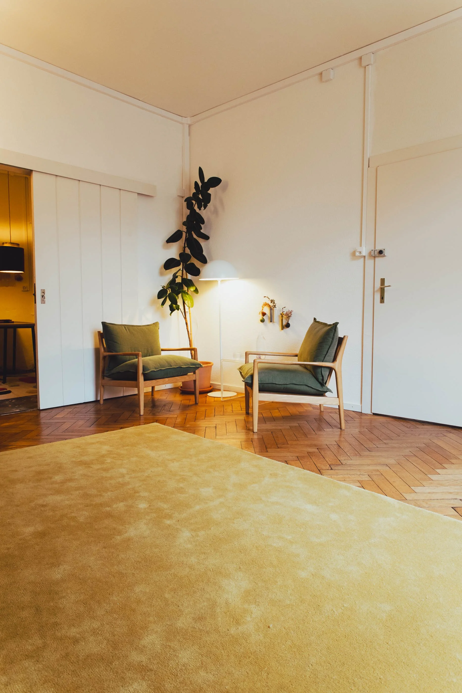 Ein stilvoll eingerichteter Praxisraum mit zwei grünen Sessel, einer Stehlampe, einer großen Zimmerpflanze und einer Holzwand.