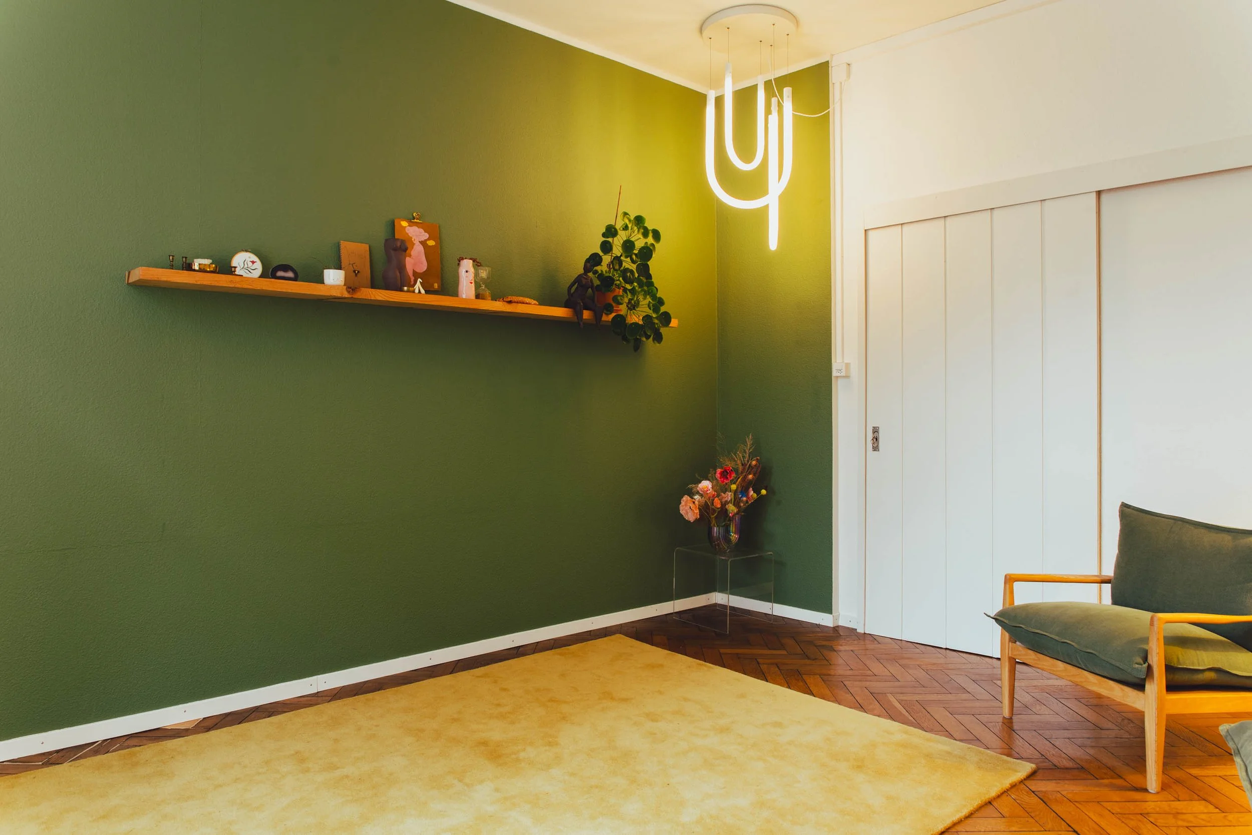 Ein Raum mit grünem Wand, Holzregal mit Dekorationen, moderner Leseleuchte, Vase mit bunten Blumen, Holzboden mit gelbem Teppich, weiße Schiebetür, Holzsessel mit grüner Polsterung.