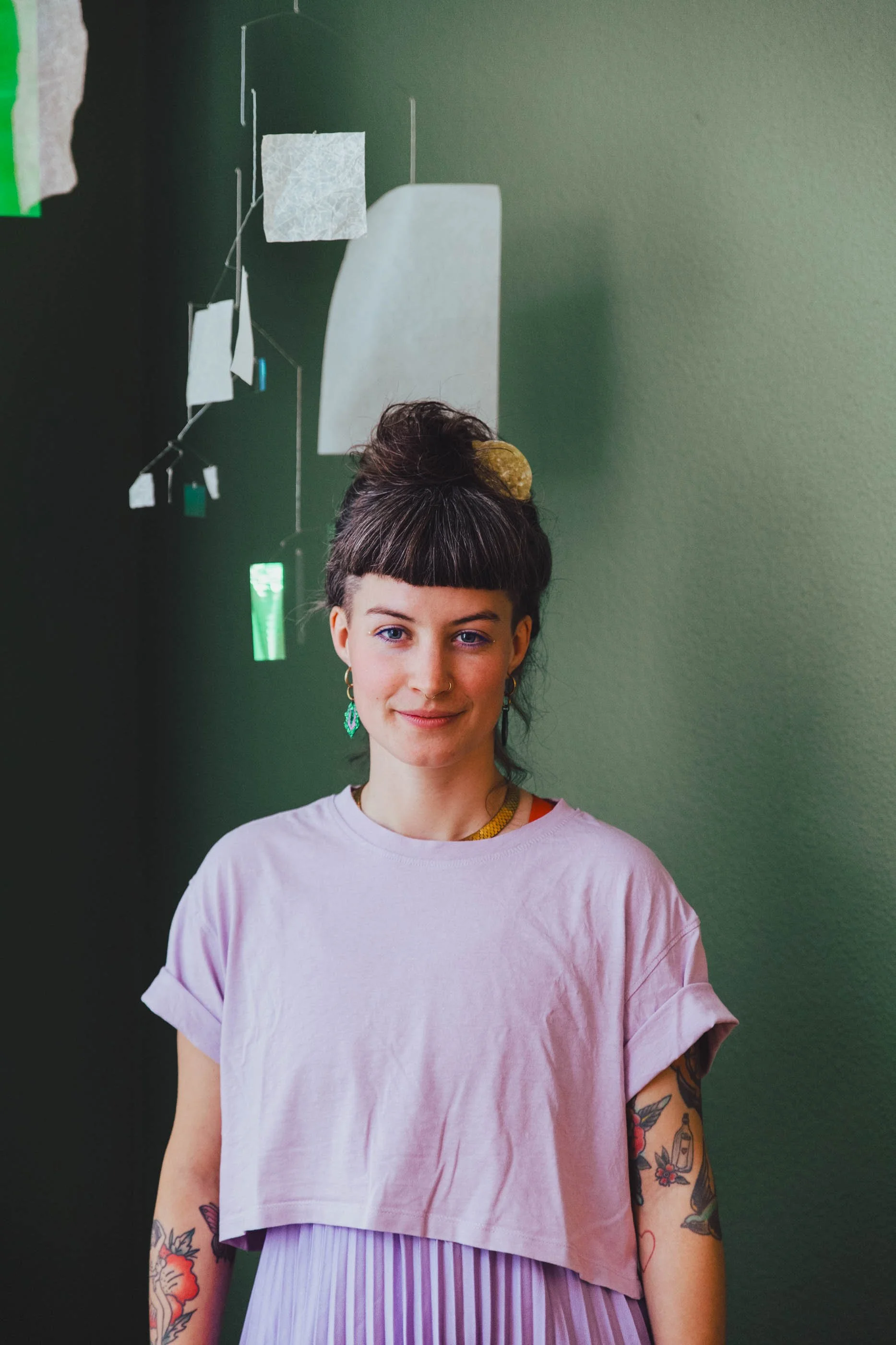 Junge Frau mit Tattoos, dunklen Haaren in Hochsteckfrisur, gepunkteter Tüllskirt, umgeben von bunten Papier- und Stoffstücken an einer grünen Wand.