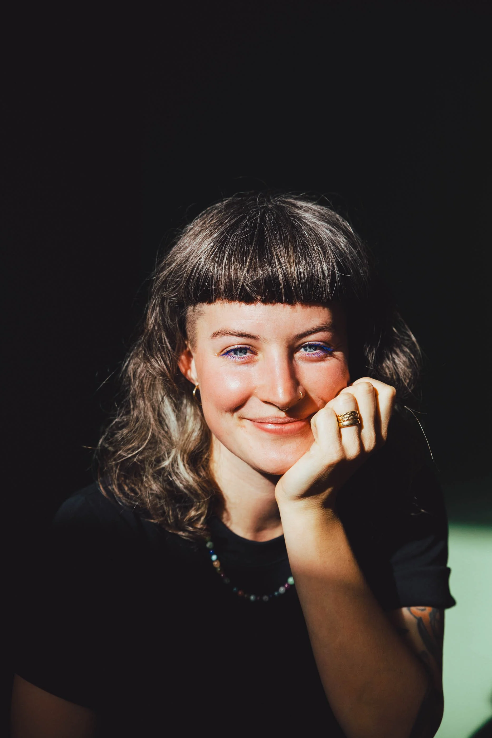 Junge Frau mit kurzen dunklen Haaren und blauen Augen, lächelt, ruht ihr Kinn auf ihrer Hand, trägt schwarze Kleidung, eine Halskette, einen Ohrring und einen Ring, vor dunklem Hintergrund.