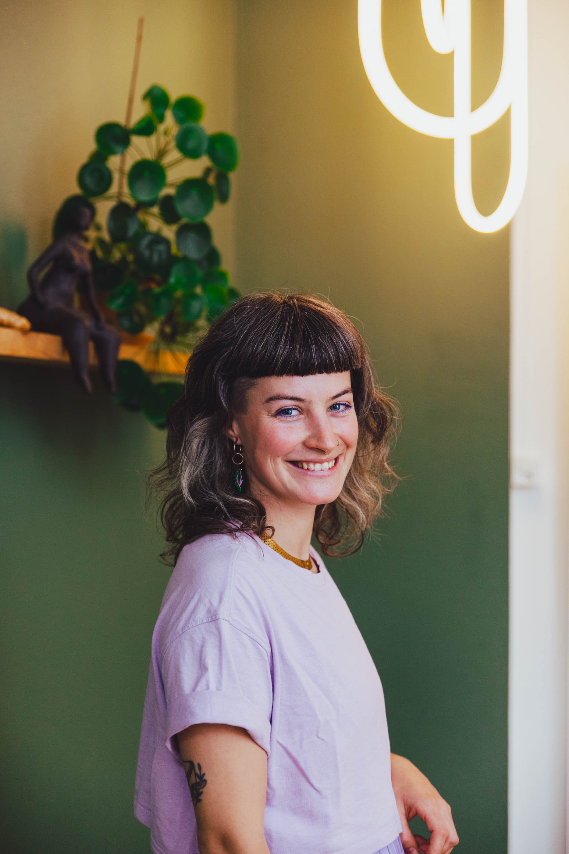 Porträt einer lachenden jungen Frau mit dunklen, gewellten Haaren, hellrosa T-Shirt, Brille und Ohrringen, vor grüner Wand mit Pflanzen und einer Neonbeleuchtung.