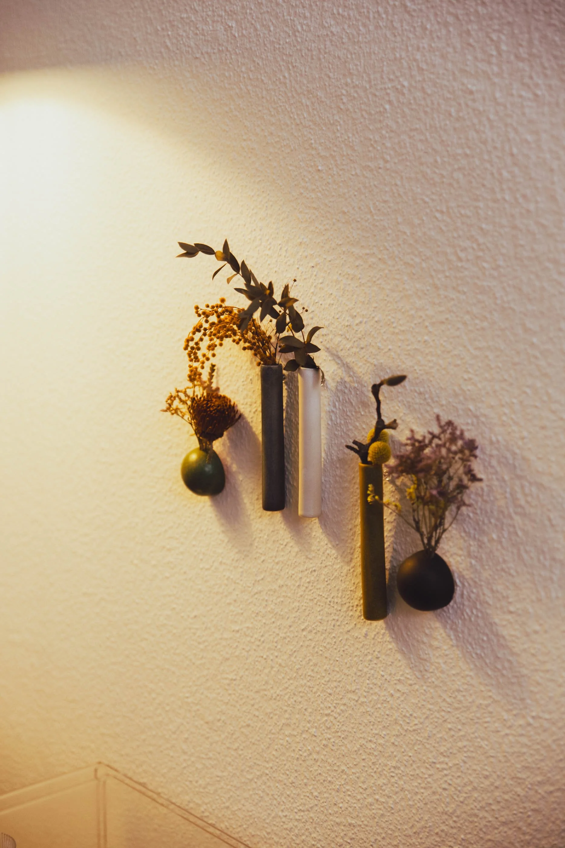 Abstrakte Wanddekoration mit mehreren kleinen Gefäßen, die jeweils mit getrockneten Blumen und Blättern gefüllt sind, an einer hellen, strukturierte Wand.