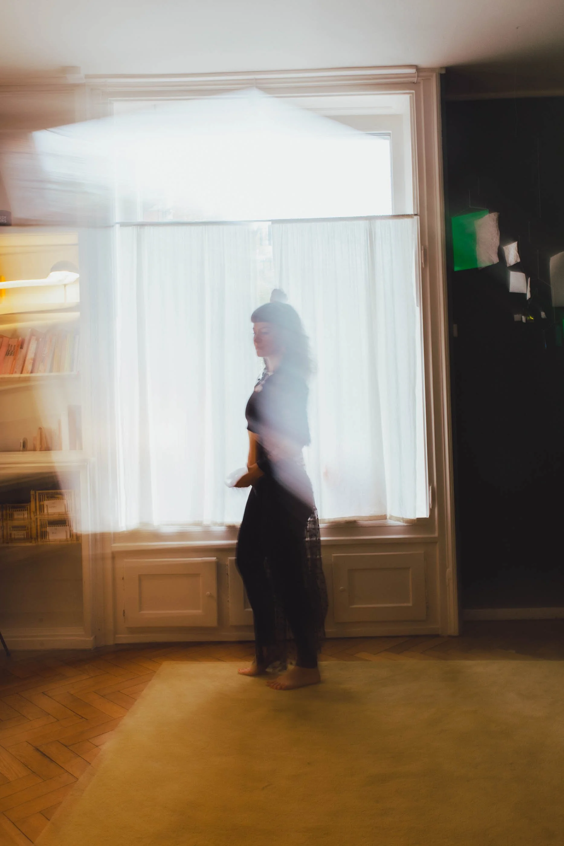 Ein Mensch steht in einem Raum vor einem großen Fenster mit weißen Vorhängen, durch das natürliches Licht einfällt. Der Mensch trägt dunkle Kleidung und ist barfuß. Die Szene wirkt durch längsverschriebene Bewegungsunschärfe.