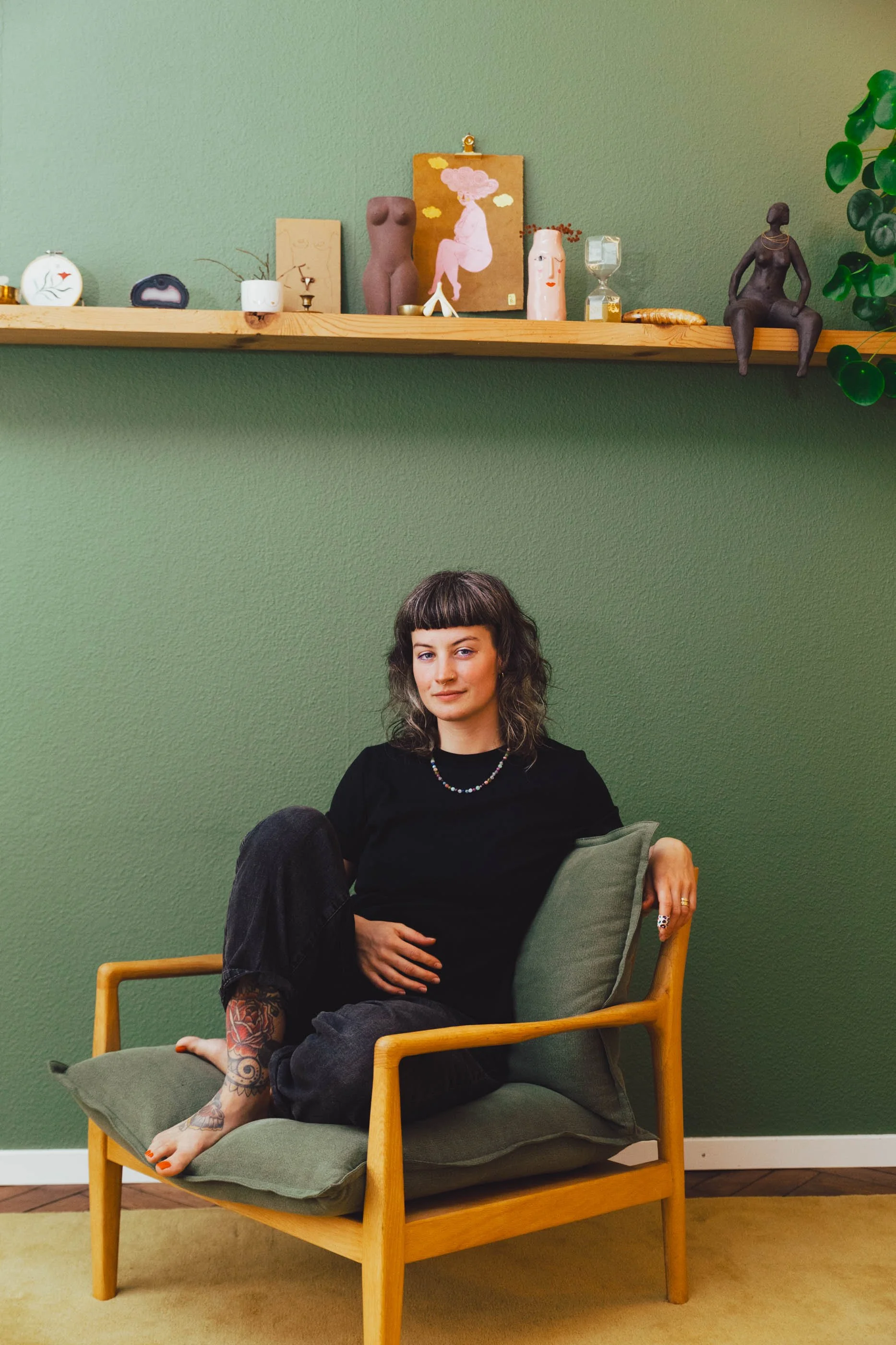 Eine junge Frau sitzt entspannt auf einem grünen Sessel vor einer grünen Wand, mit einer Vielzahl von Dekorationsgegenständen und Kunstwerken auf einem Holzregal darüber.