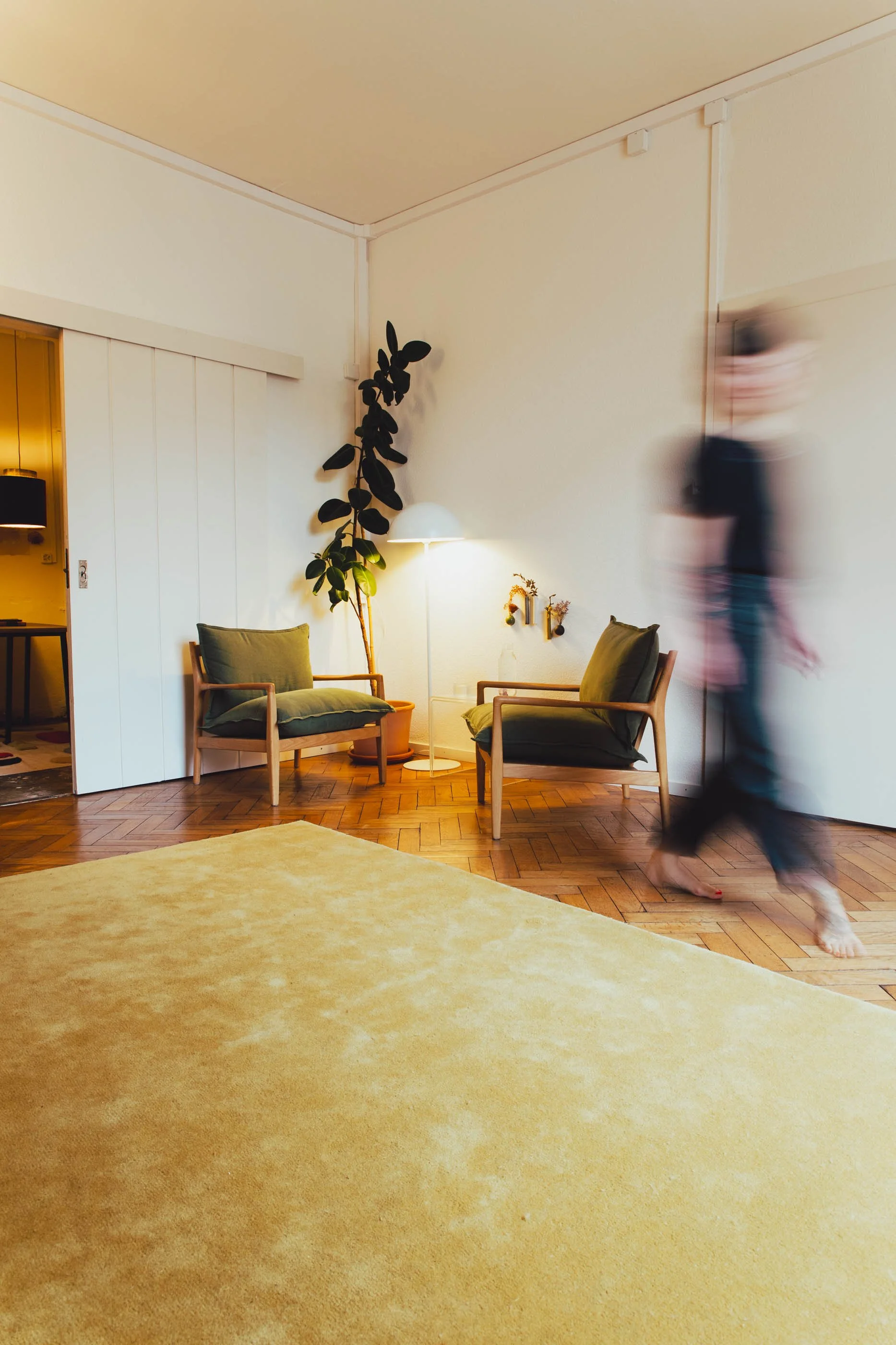 Ein Wohnzimmer mit zwei grünen Sesseln, einem stehenden Lampenschirm, einer Pflanze, Holzfußboden, gelbem Teppich und einer Person, die schnell vorbeigeht.