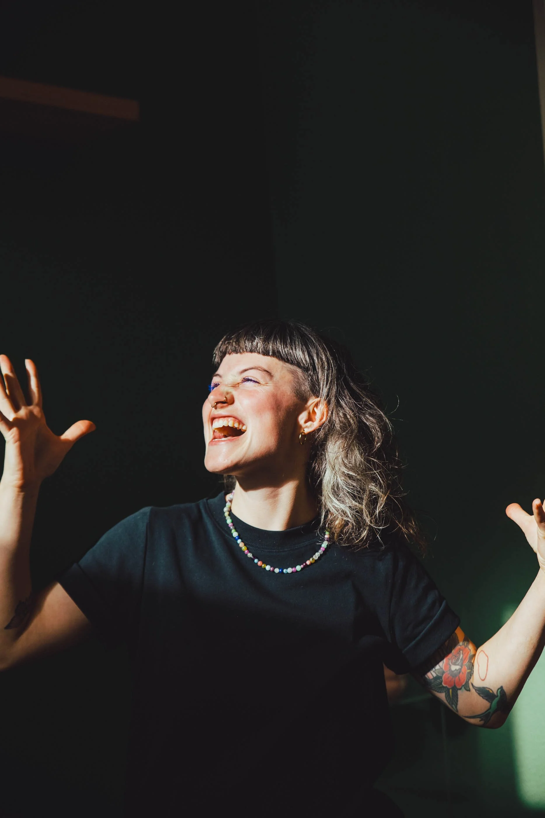 Freudige junge Frau mit Tattoos, buntem Schmuck und kurzem Fransenhaarschnitt, die lachen und mit den Armen in der Luft gestikuliert.