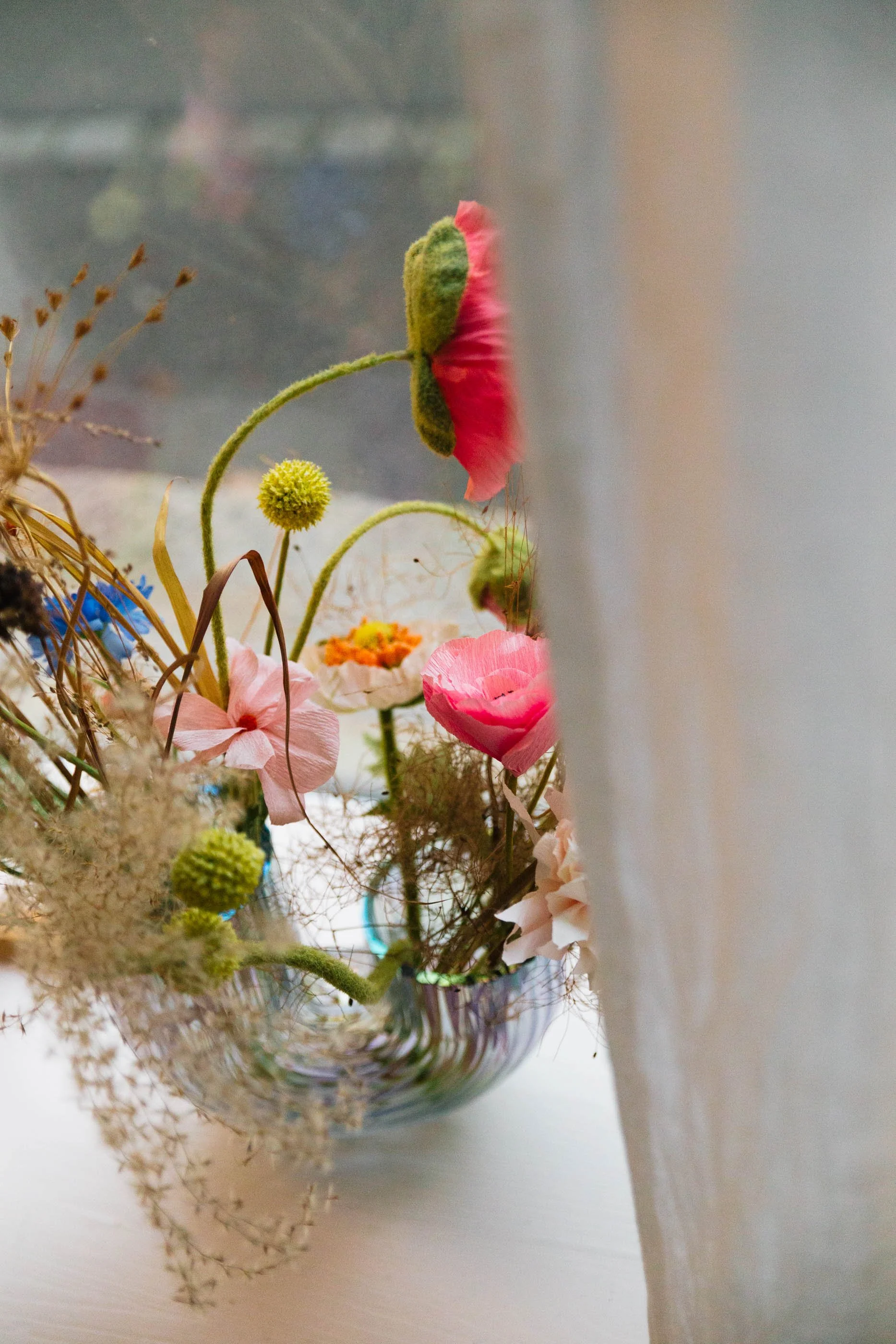 Bunte Blumen in einer Glaskaraffe, halb verdeckt durch einen weißen Vorhang