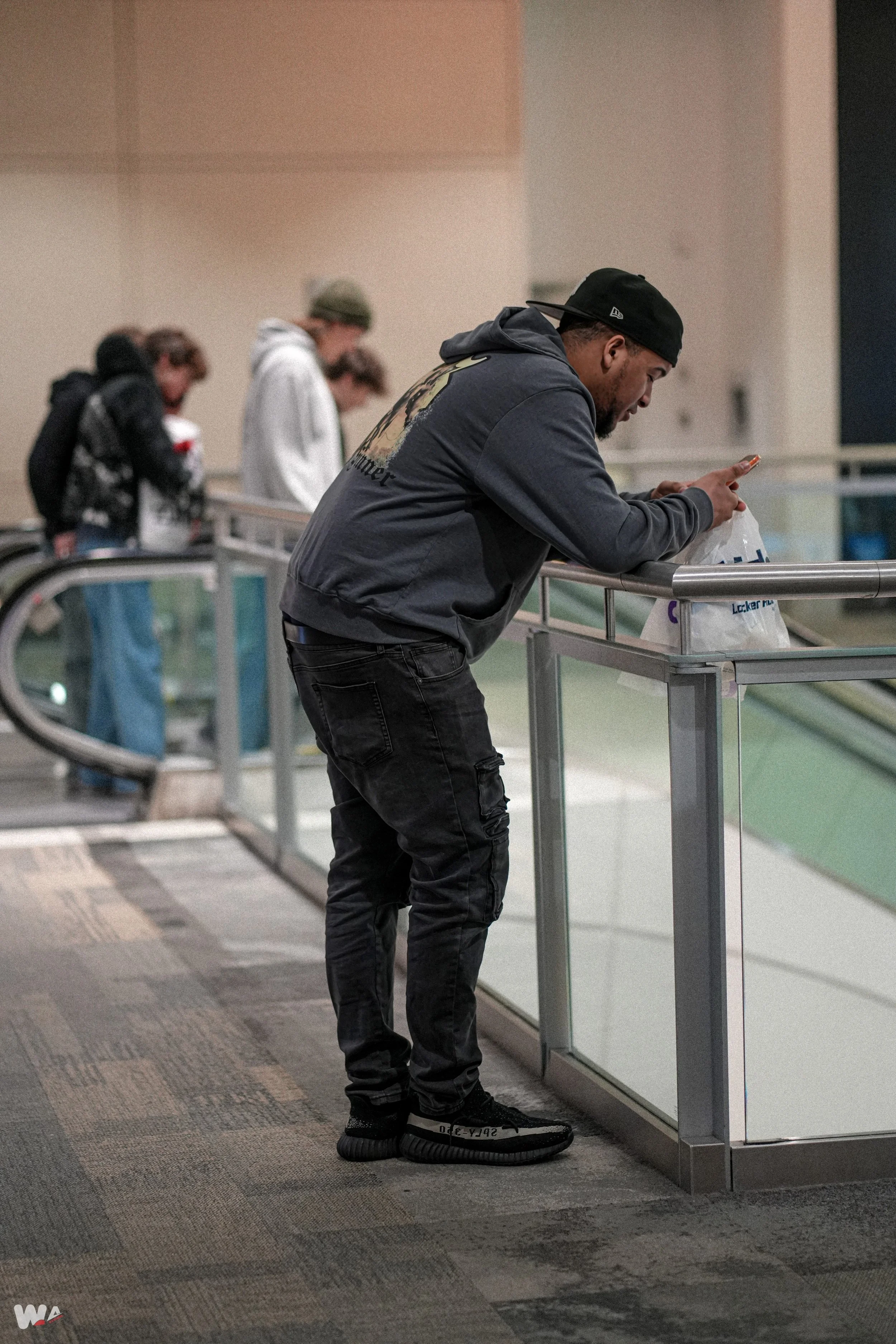 Man leaning on railing at airport, looking at phone, with people in line behind him.