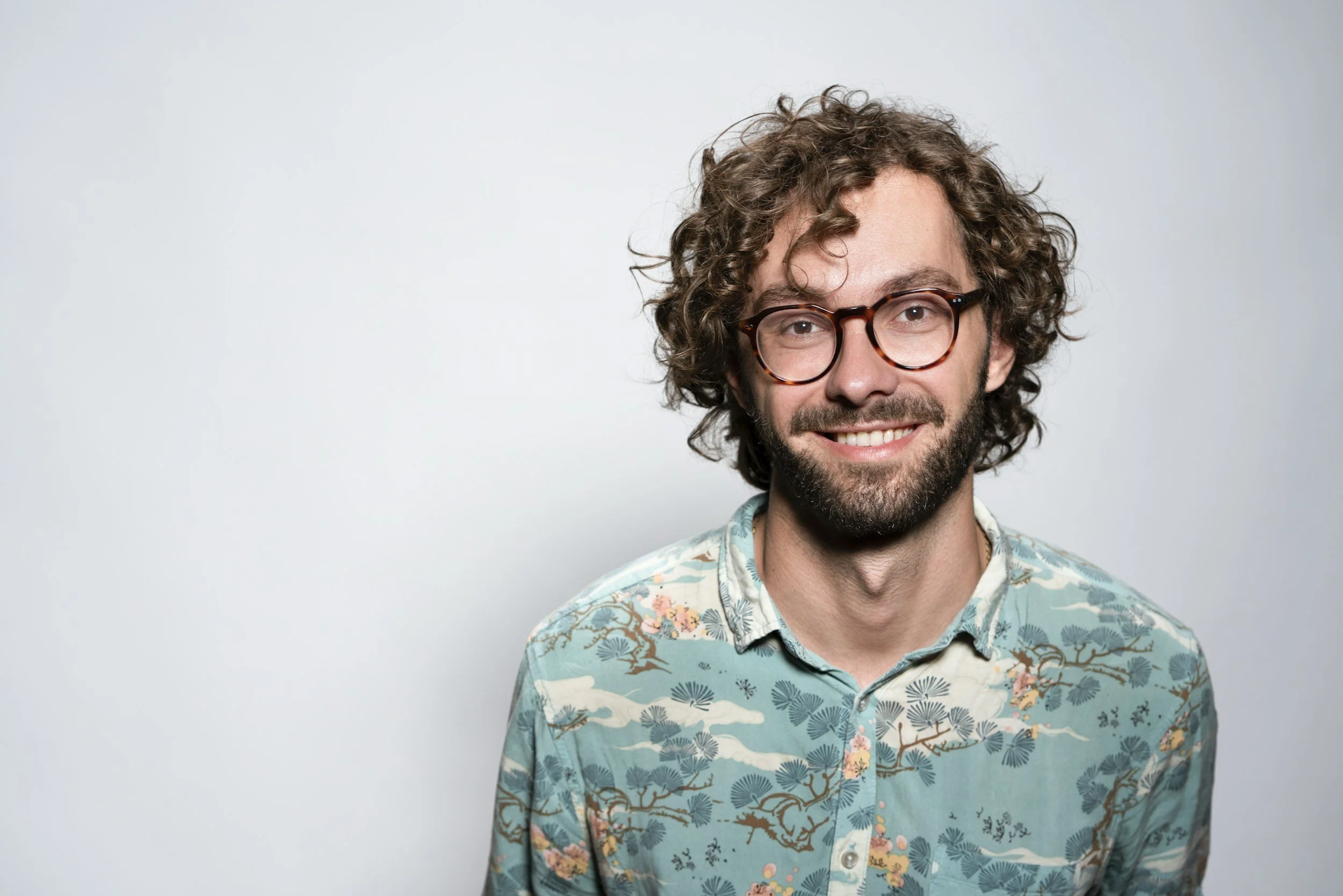 A man with curly hair, glasses, and a beard, smiling while wearing a colorful patterned shirt against a plain light gray background.