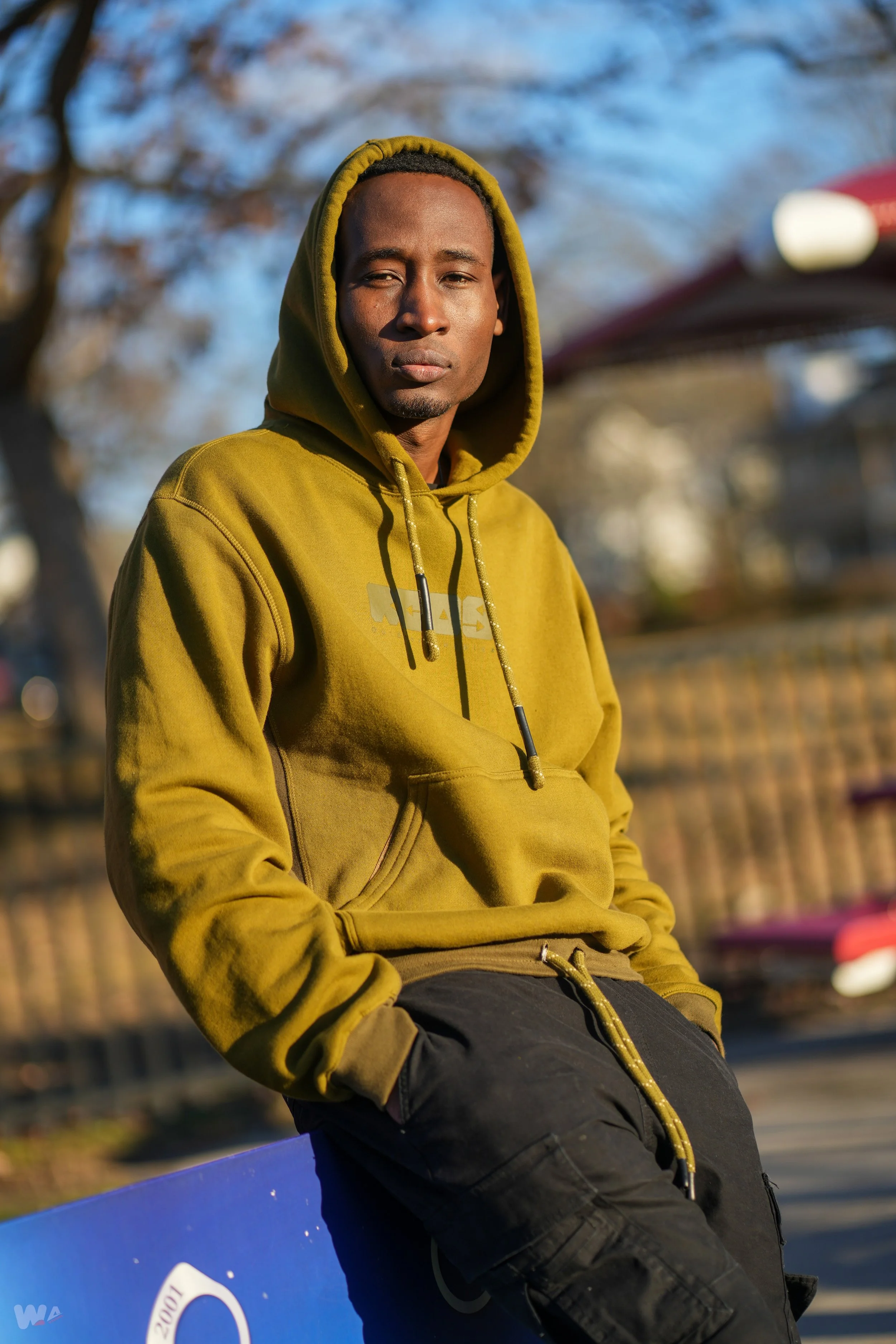 A man wearing a yellow hoodie and black pants, sitting outdoors on a blue surface, with a blurred background of trees and a fence.