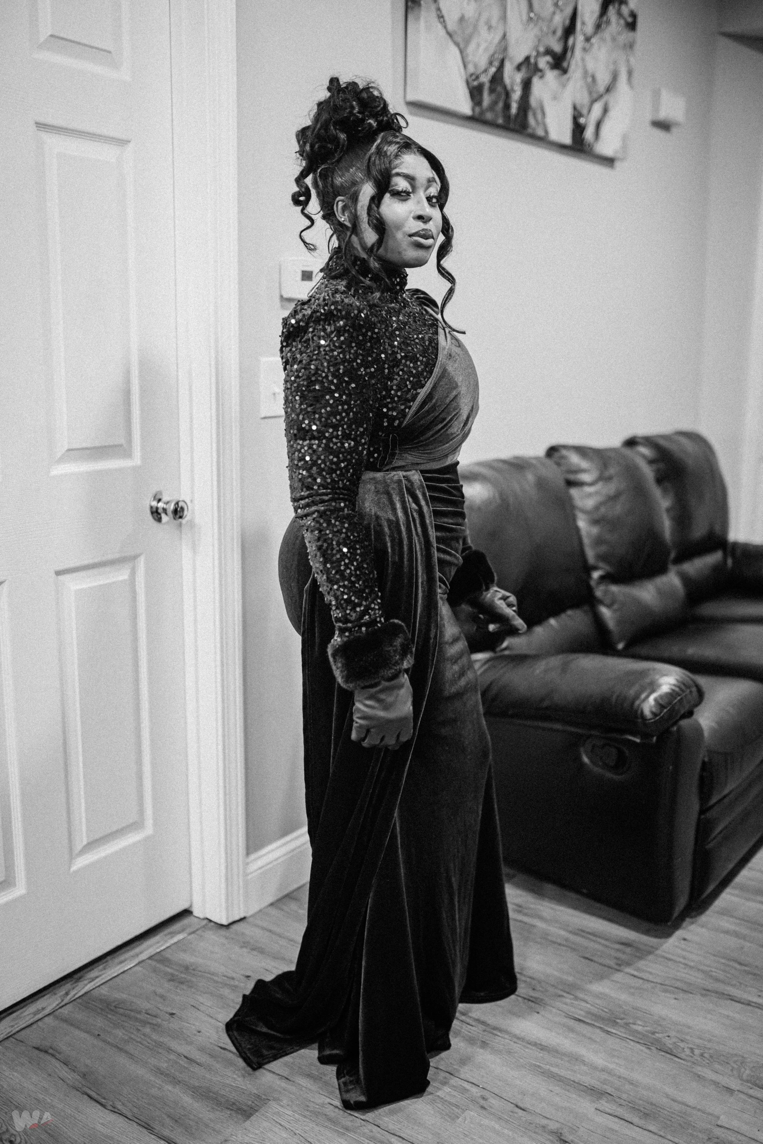 A woman standing indoors in front of a leather couch, dressed in a glamorous, sequined top and velvet pants, with styled hair and makeup.