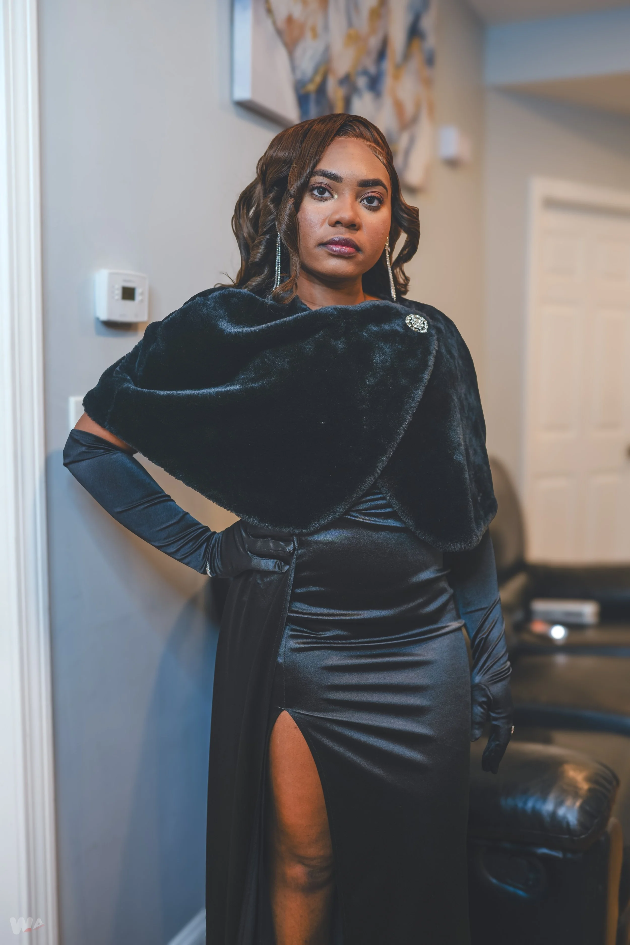 A woman dressed in a long, black, elegant gown with a high slit, wearing long black gloves and a black fur cape, standing indoors with a neutral expression.