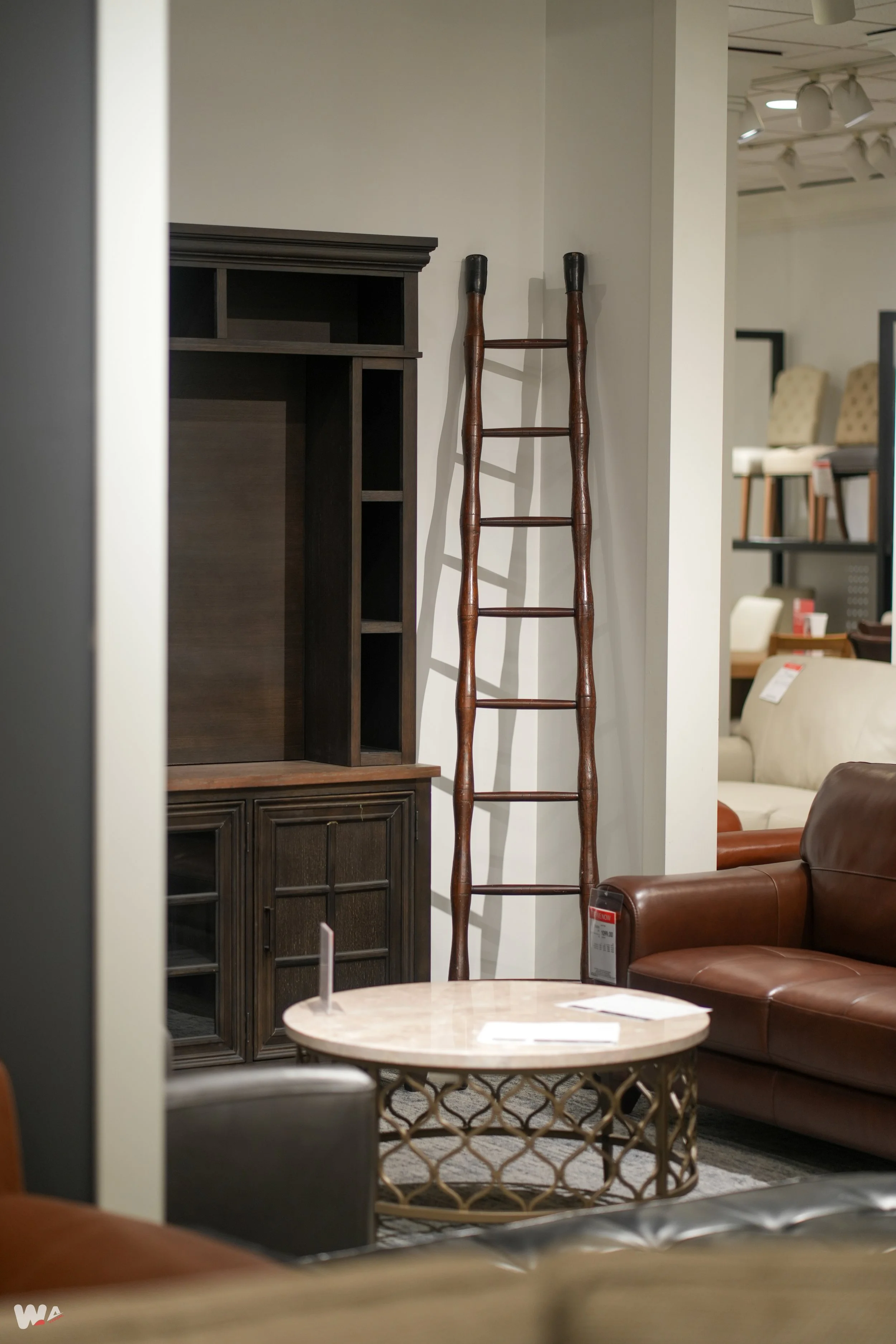 Inside a furniture store, a wooden ladder with black tips leaning against a white wall, with other furniture items like a dark wooden TV stand, a round marble-top coffee table, and leather sofas visible around.