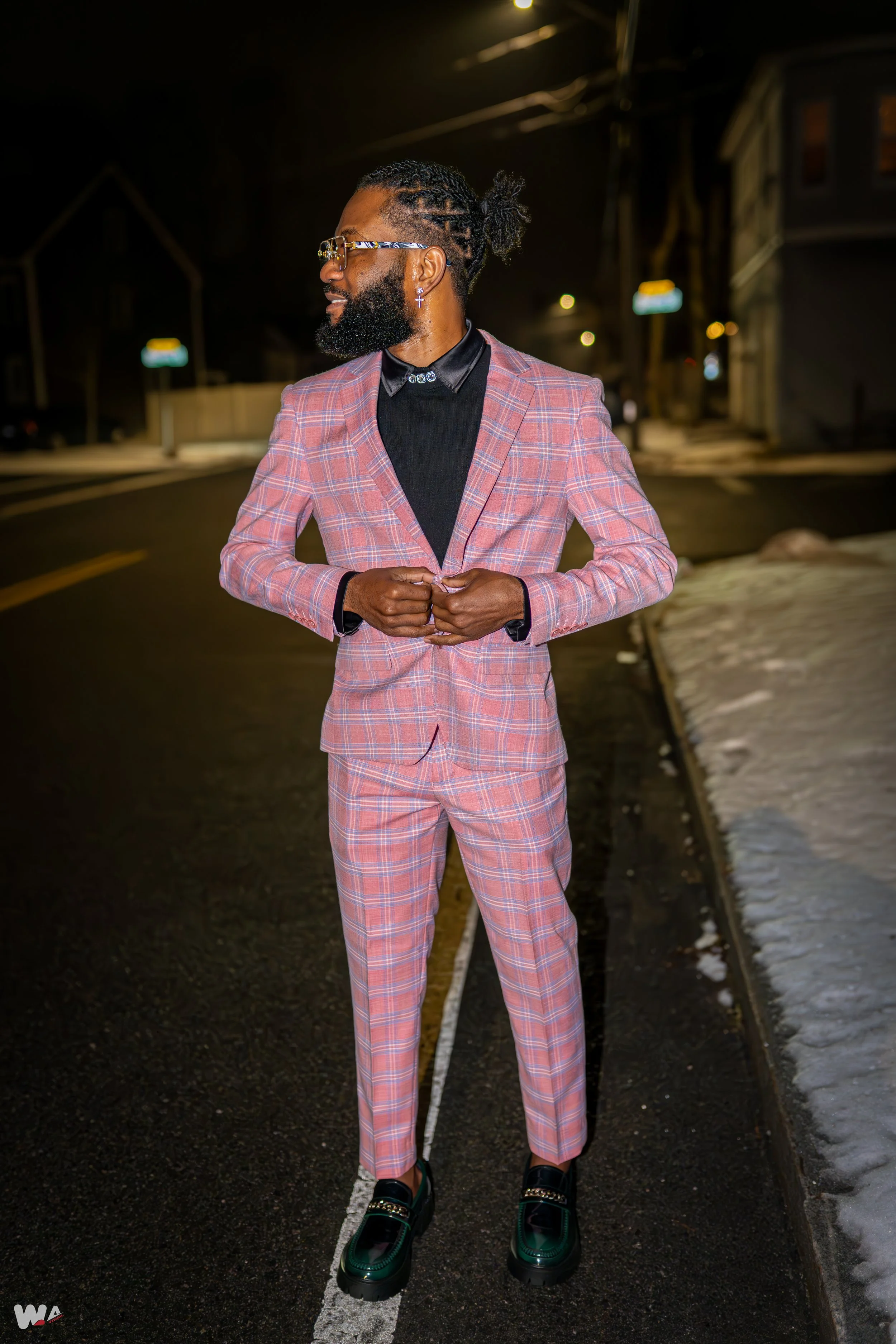 A man dressed in a pink plaid suit and black shirt standing on a dark street at night.