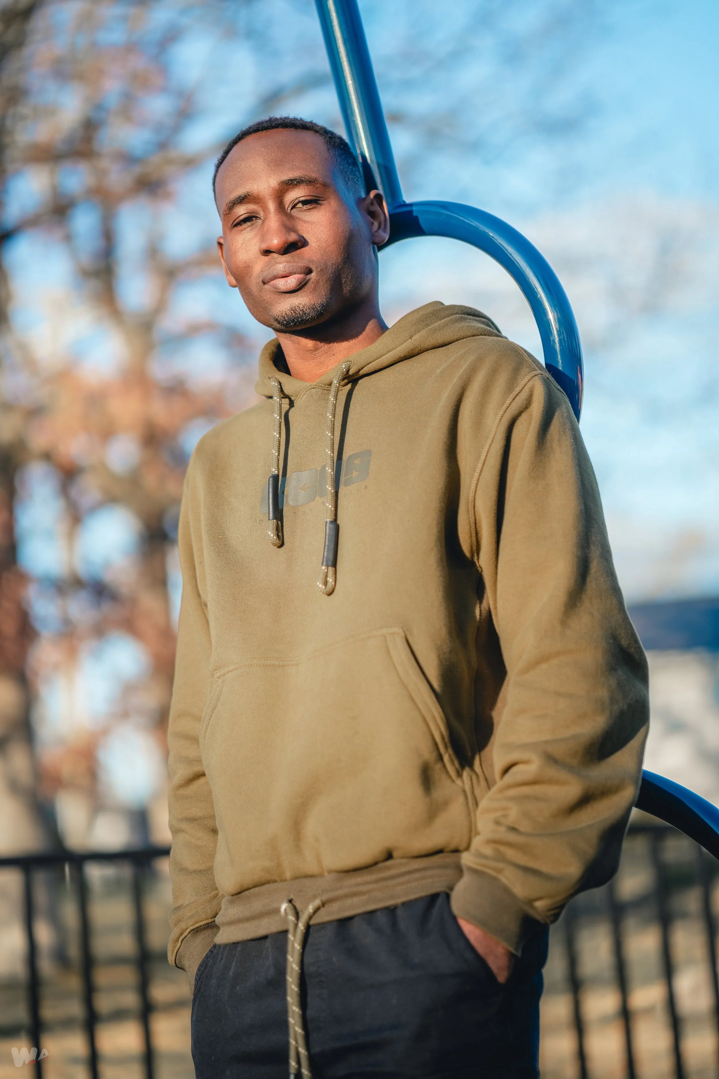 A young man in a khaki hoodie with drawstrings stands outdoors near a playground, with a blue metal structure behind him. He has short hair and is looking at the camera with a neutral expression. Blurred trees are visible in the background, indicatin