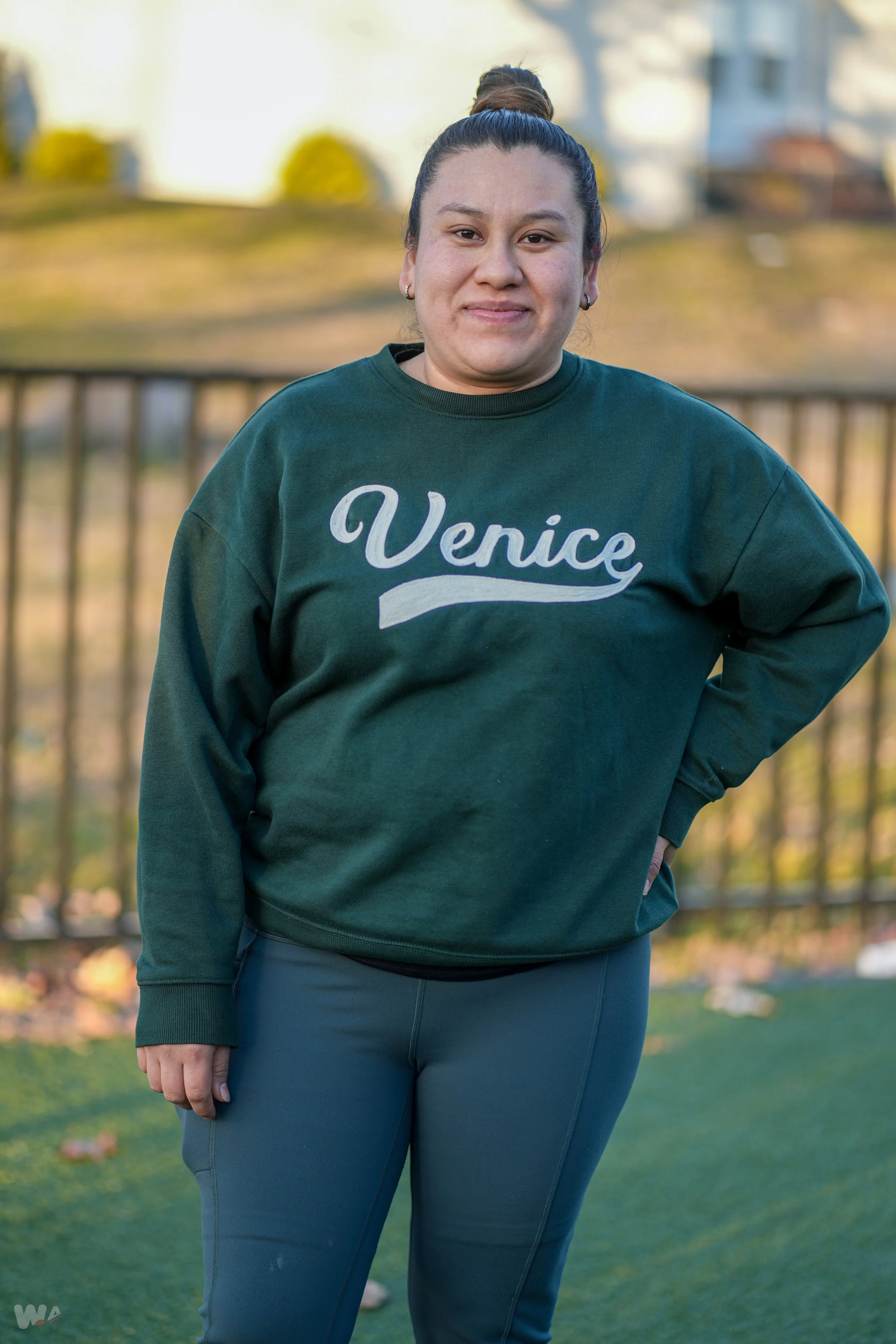 A woman with dark hair tied in a bun, wearing a green hoodie with 'Venice' written on it and blue pants, standing outdoors with a blurred background of trees and a railing.