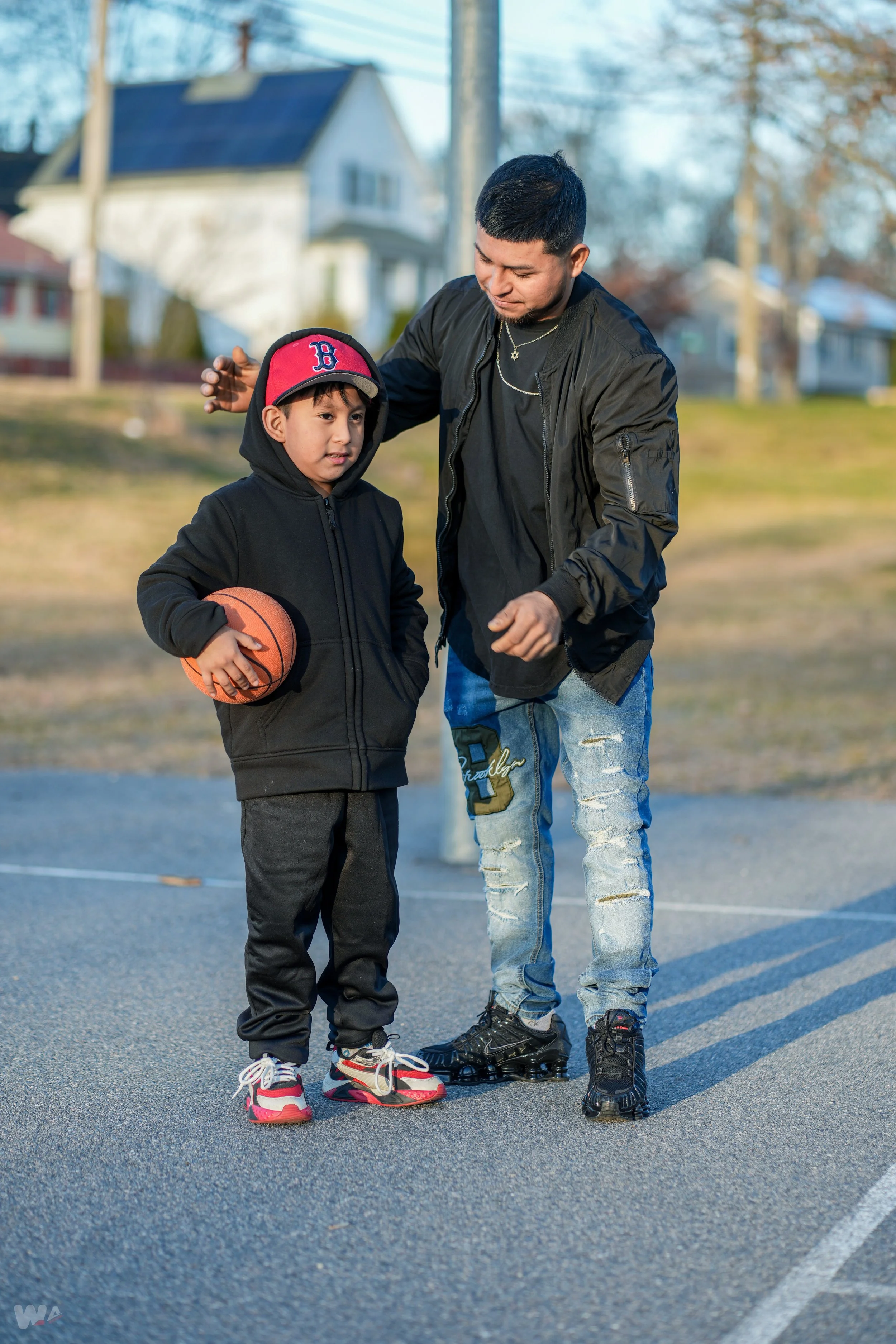 A young boy holding a basketball stands next to an adult man on a basketball court outside, with houses and trees in the background. The man appears to be coaching or giving instructions to the boy.