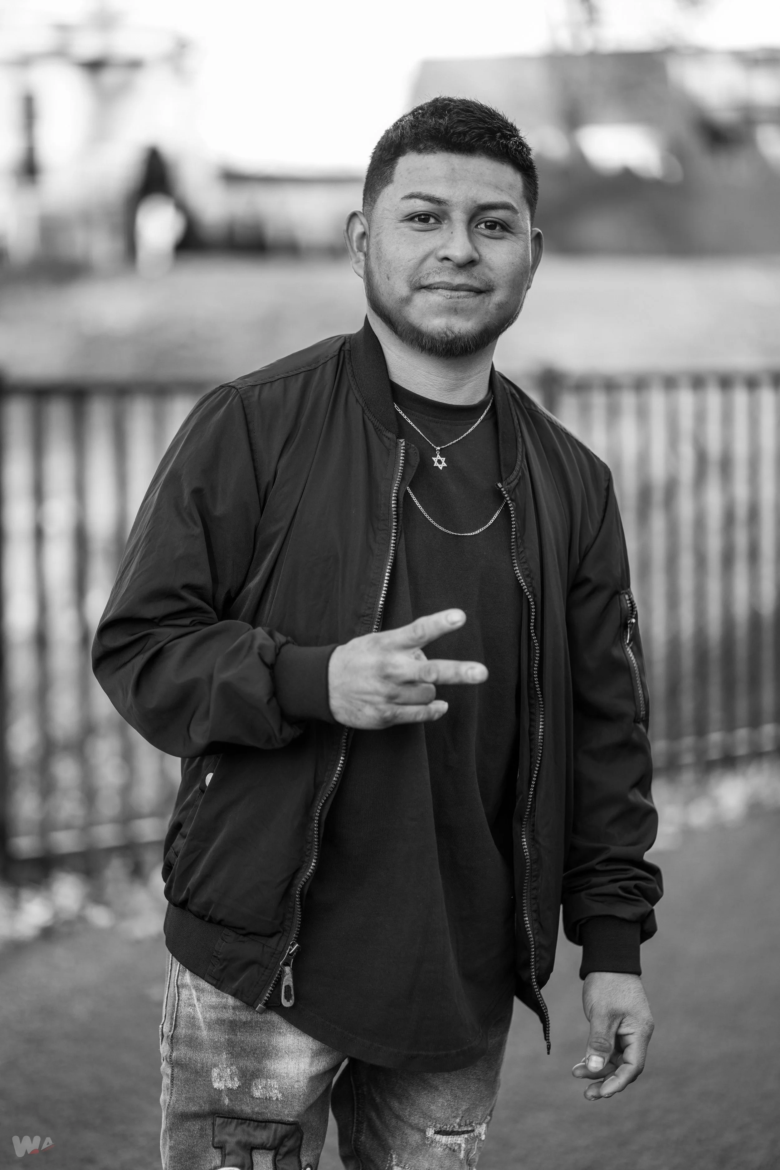 A young man with short hair and a goatee, wearing a black jacket and t-shirt, making a peace sign with his right hand, standing outdoors near a railing with blurred boats in the background.