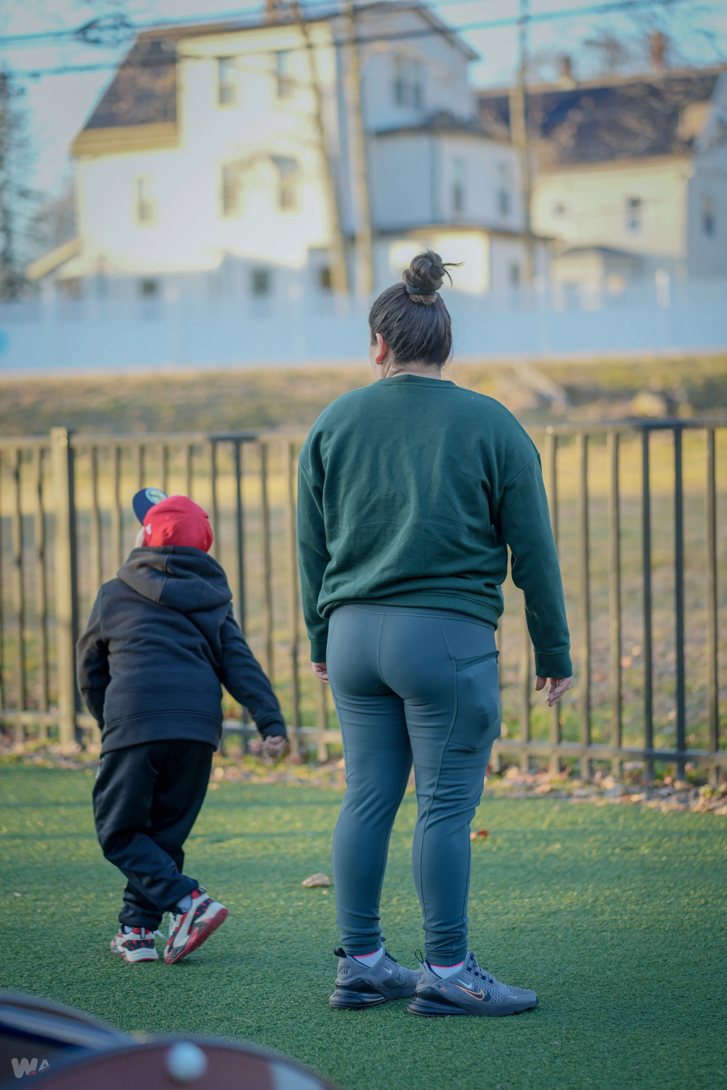 A woman and a child walking on a grassy field near a black fence, with houses and trees in the background on a clear day.
