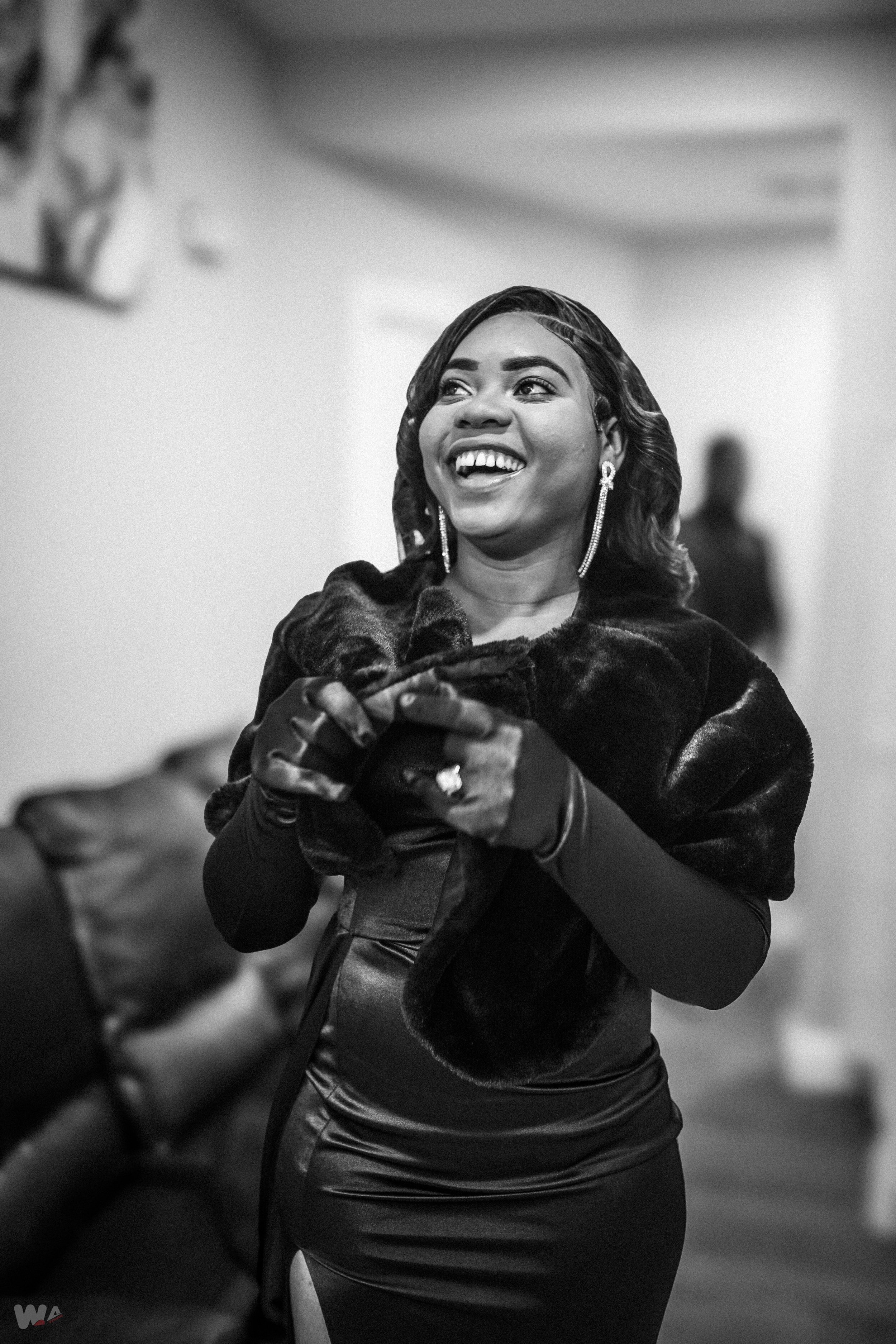 A smiling woman in a black dress and faux fur stole, wearing earrings and rings, standing indoors with a person blurry in the background.
