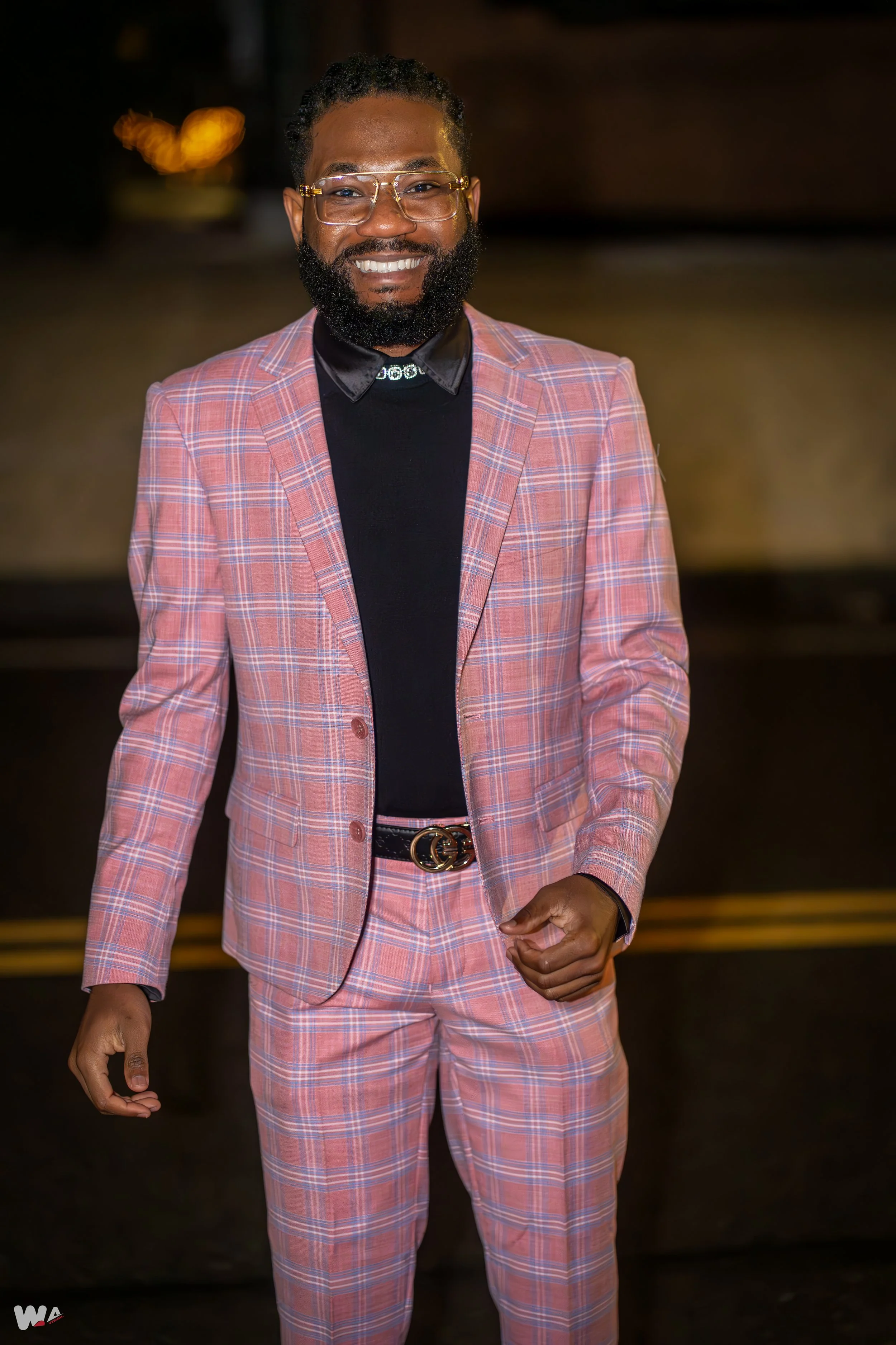 A smiling man with glasses, a beard, and short curly hair, wearing a pink plaid suit with a black shirt and a Gucci belt, standing indoors with a blurred background.