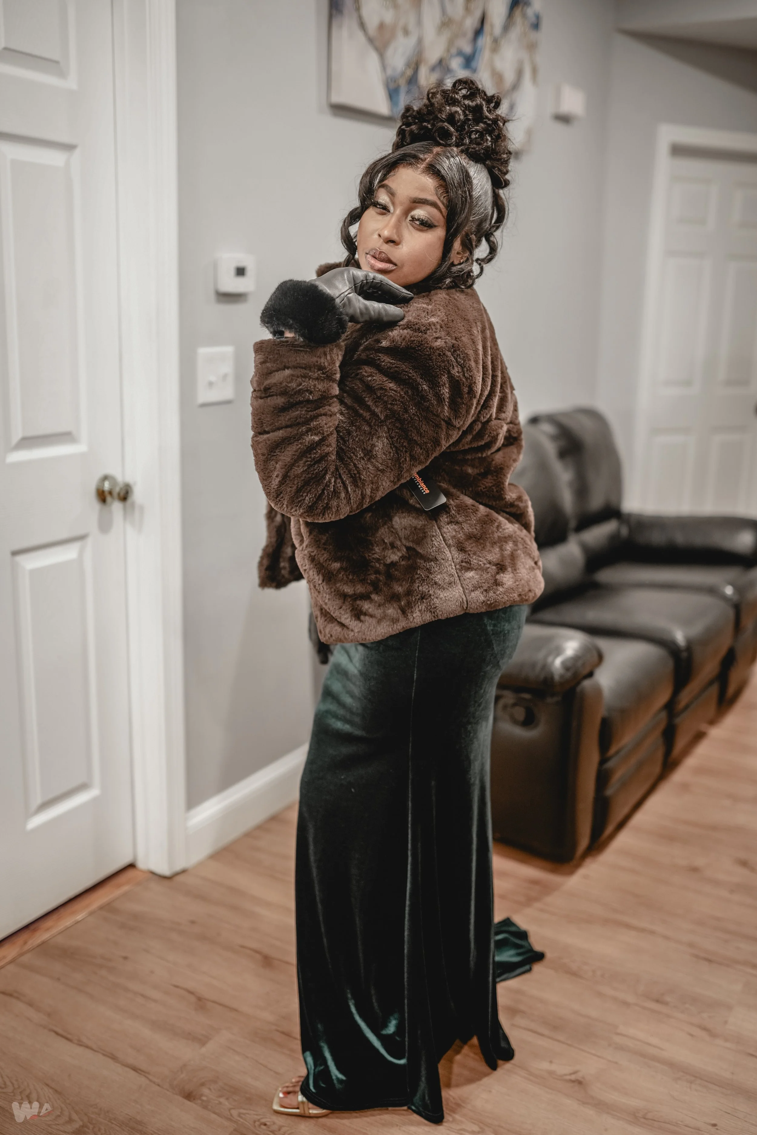 A woman standing inside a house, wearing a brown fur coat, black velvet pants, and black gloves, striking a pose with her hand near her face.