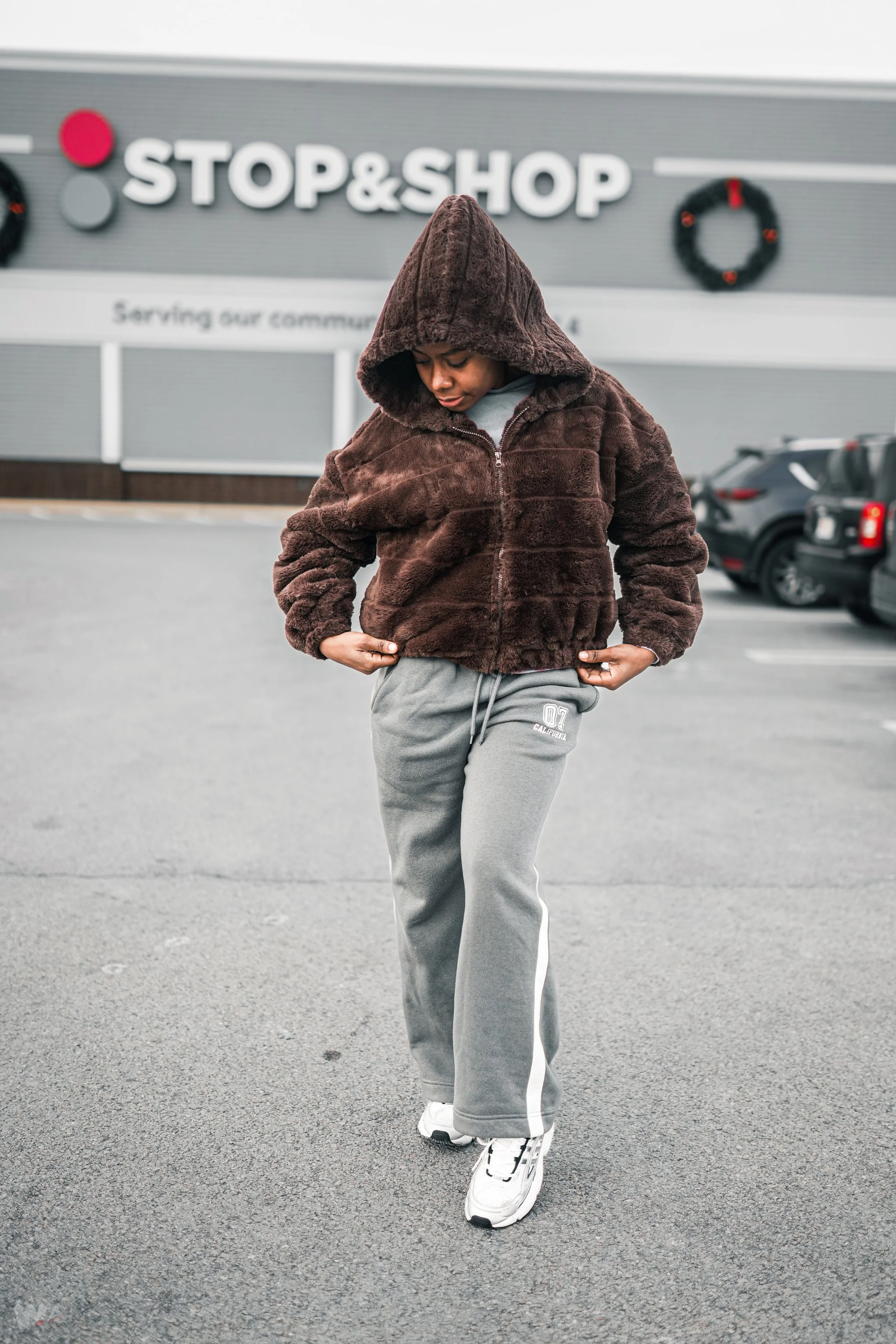 A person in a brown fuzzy hoodie, gray sweatpants, and white sneakers standing in a parking lot in front of a store with a sign that reads 'STOP & SHOP'.
