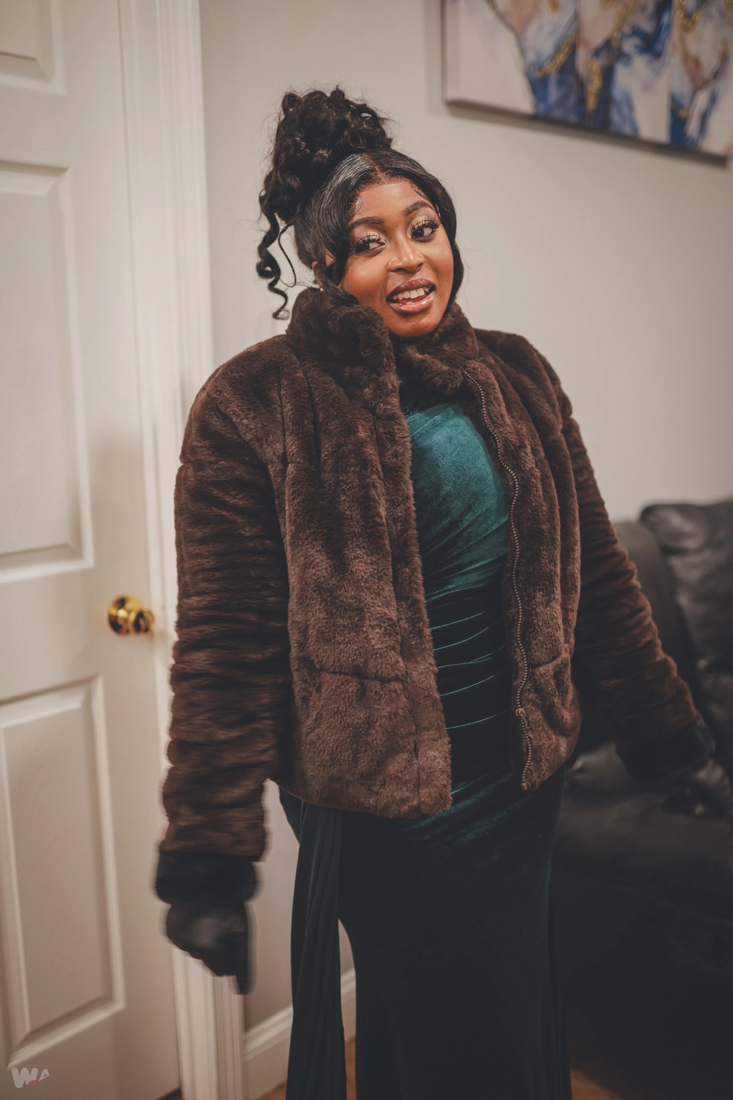 A woman with dark hair in a curly updo, wearing a brown faux fur jacket, a dark green velvet top, and a long dark skirt, standing in a living room with a white door and a black leather couch.