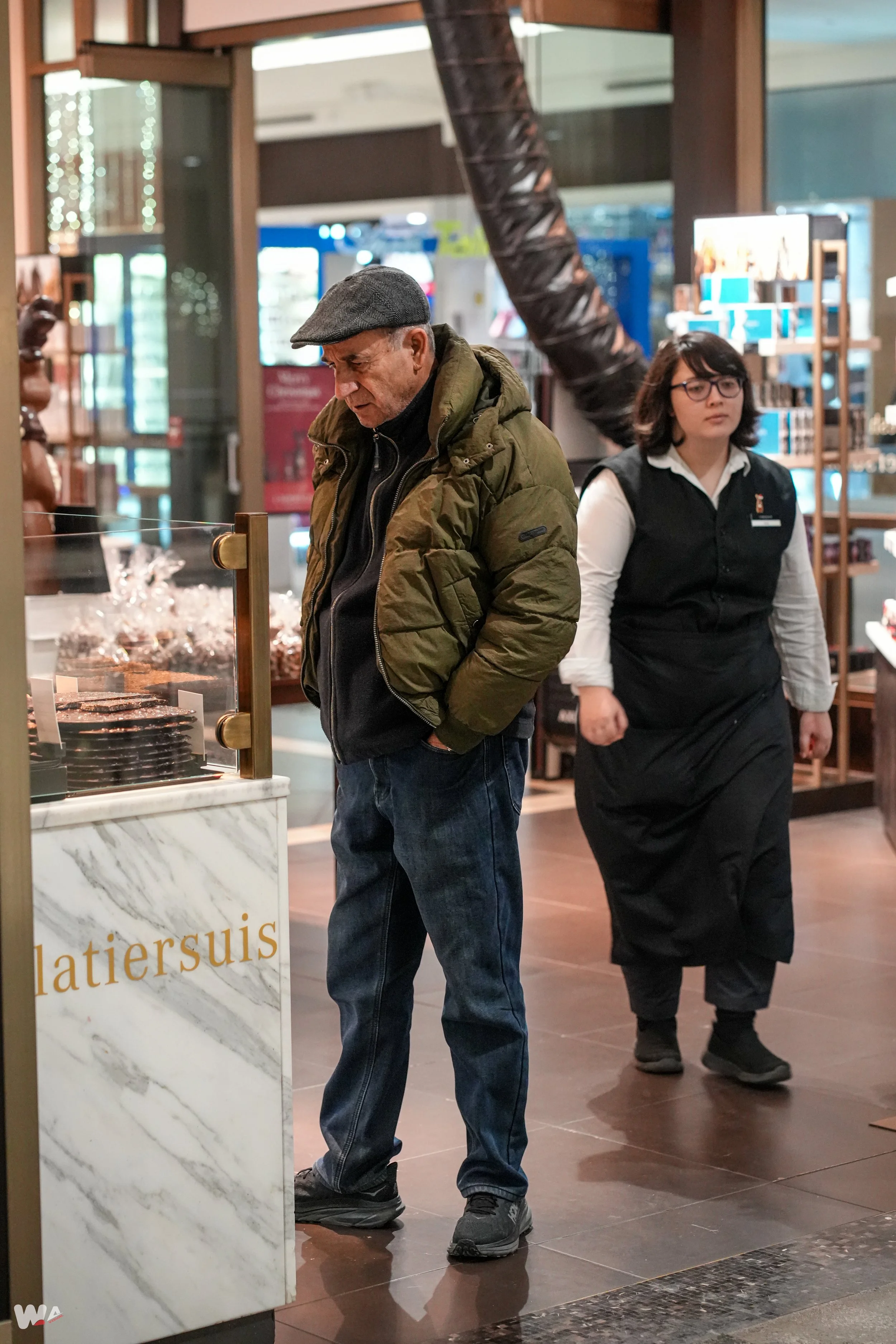 An elderly man in a green jacket and gray cap inside a store, looking at a display case. A young woman dressed as a store employee walks past in the background.