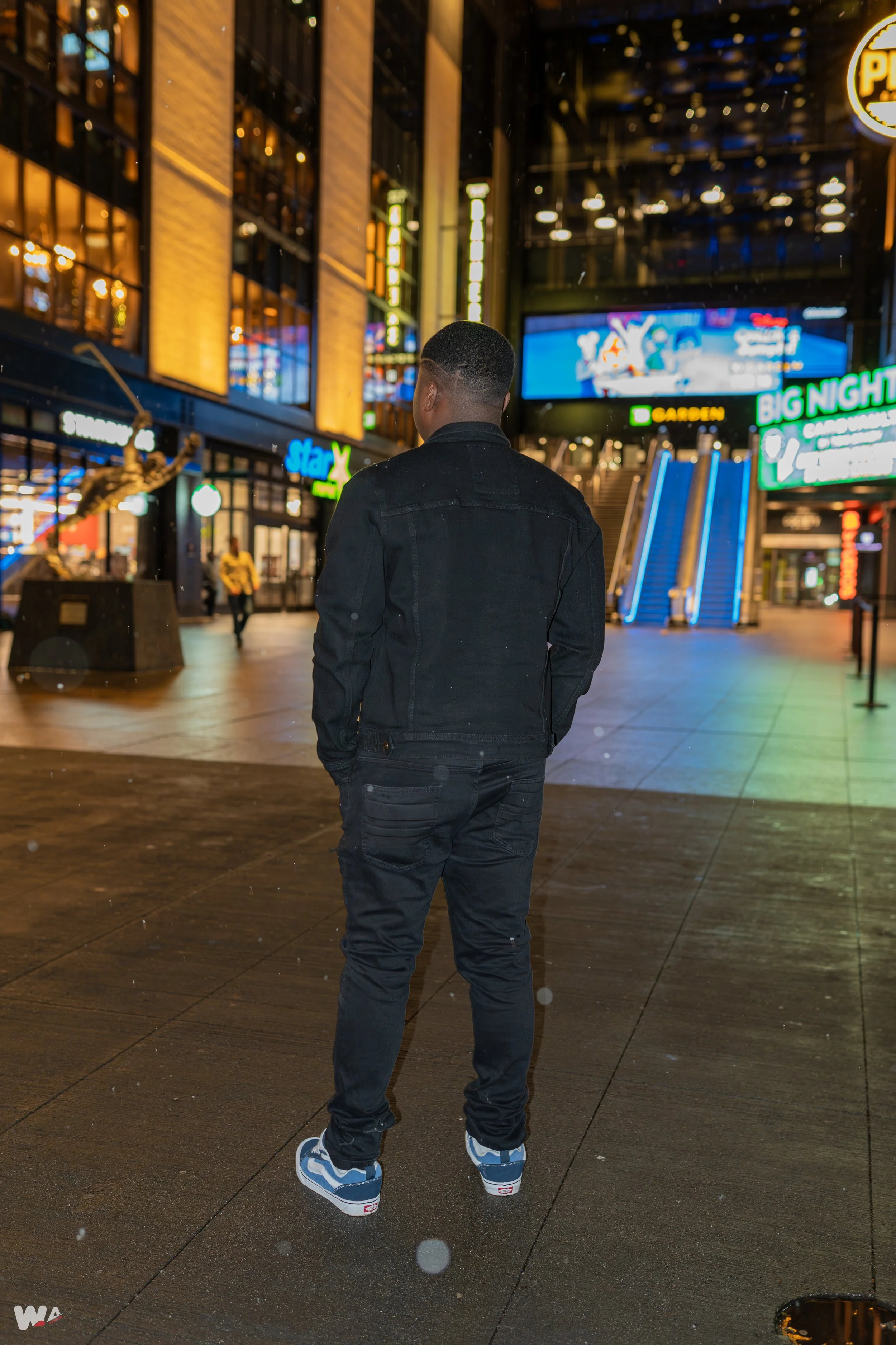 A young man dressed in black standing in an illuminated urban shopping area at night, with neon signs and escalators in the background.