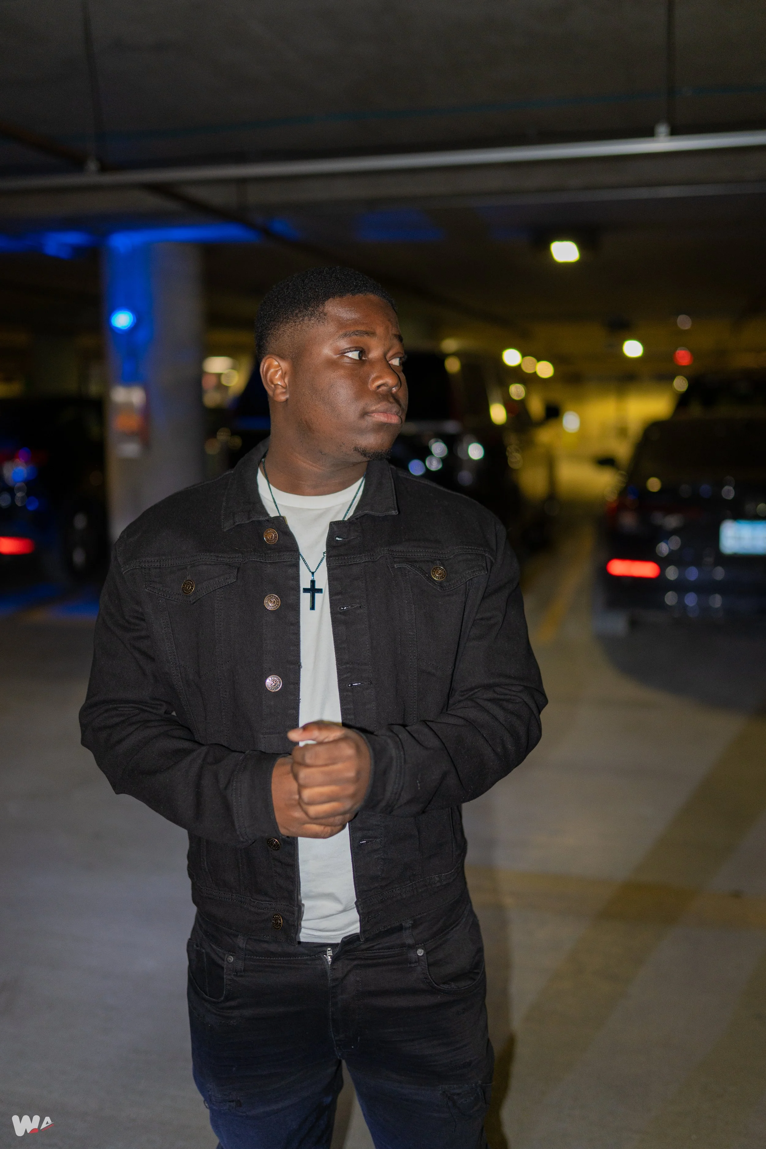 A young man in a black jacket and white shirt with a cross necklace stands in a parking garage at night, looking to the side. Cars are visible in the background.