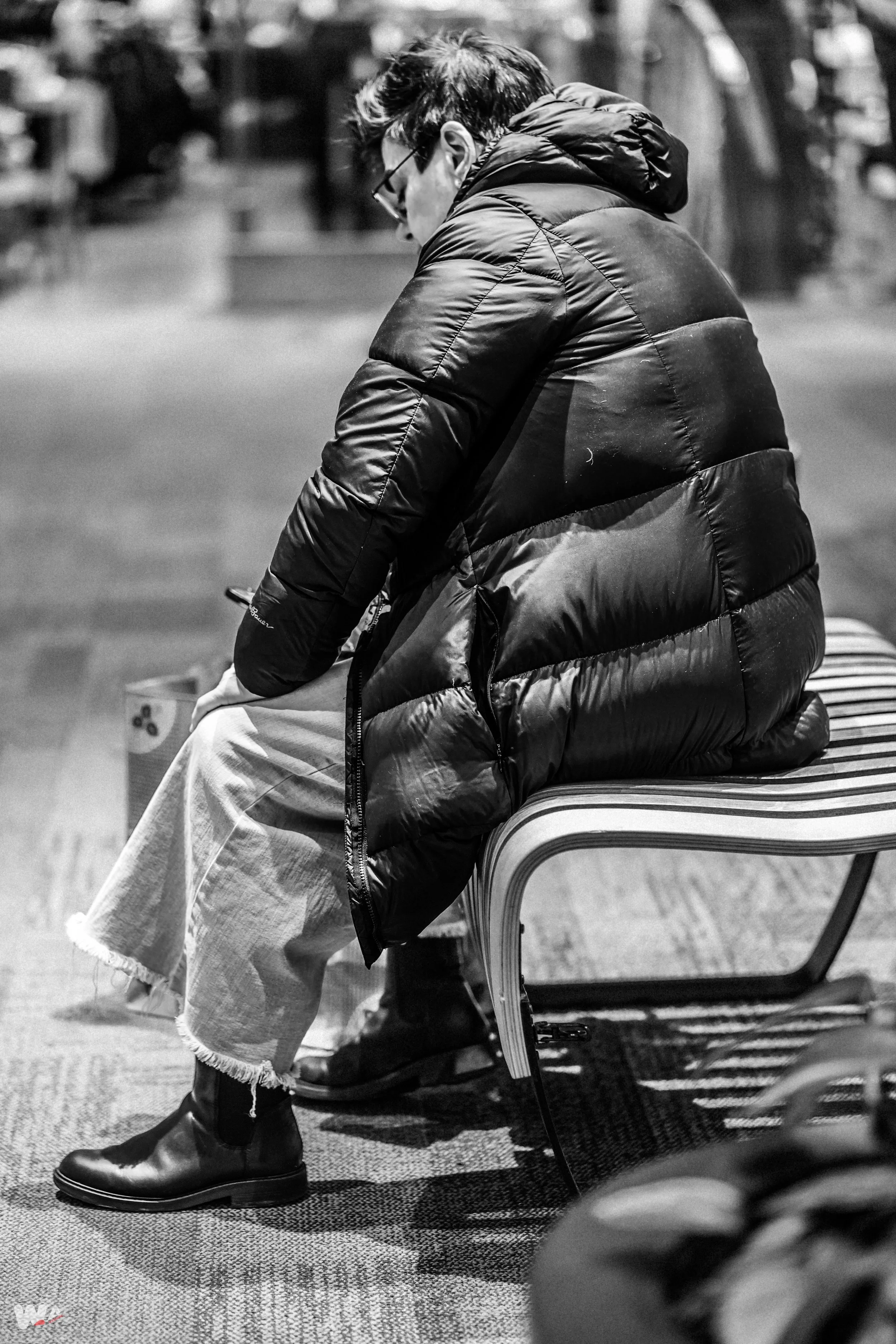 A person wearing glasses and a puffy jacket sitting on a striped bench, looking at a phone in a public indoor space.