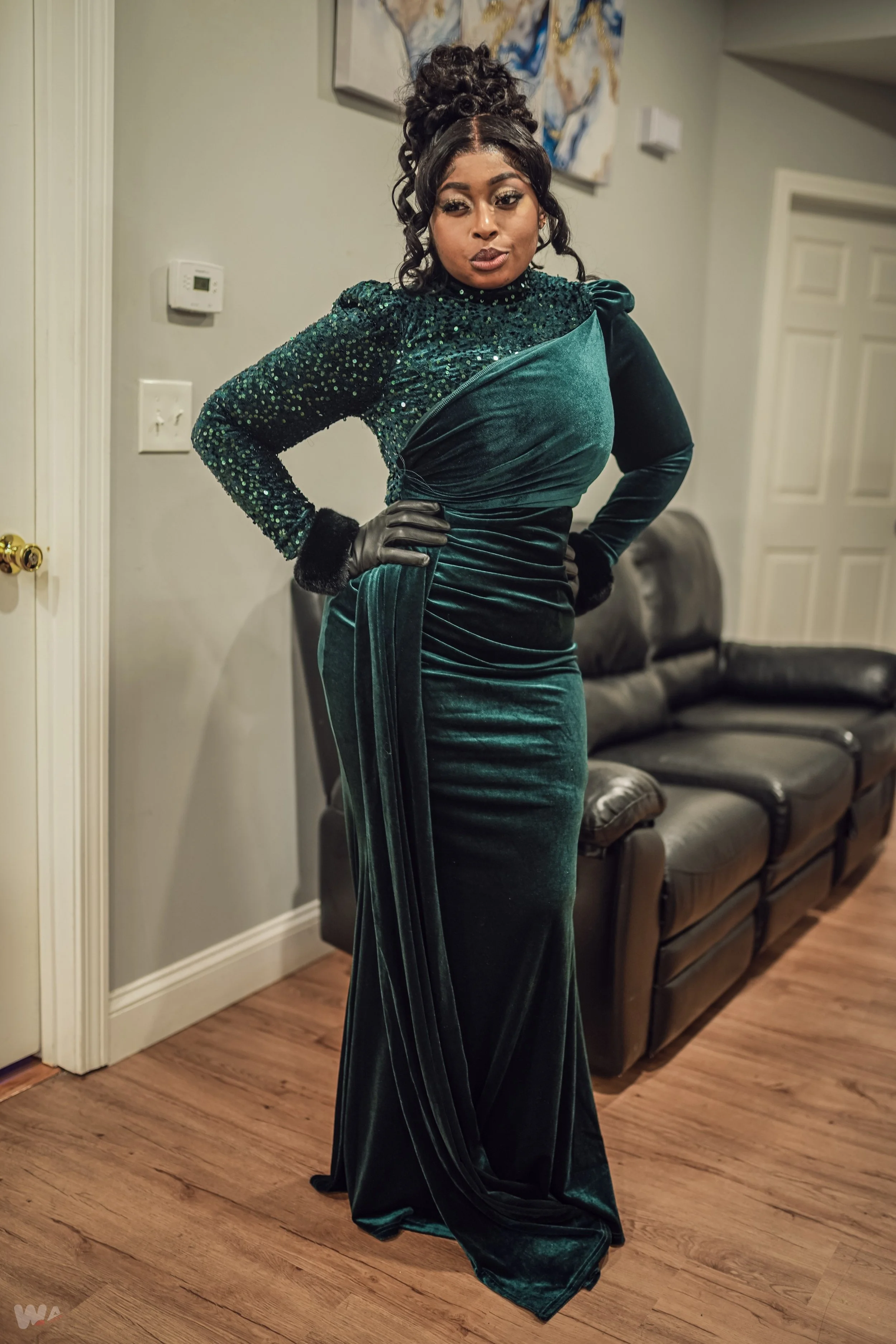 A woman wearing an elegant, dark green velvet gown with long sleeves, a high neckline, and a draped design, standing inside a room with hardwood floors and a black leather couch.