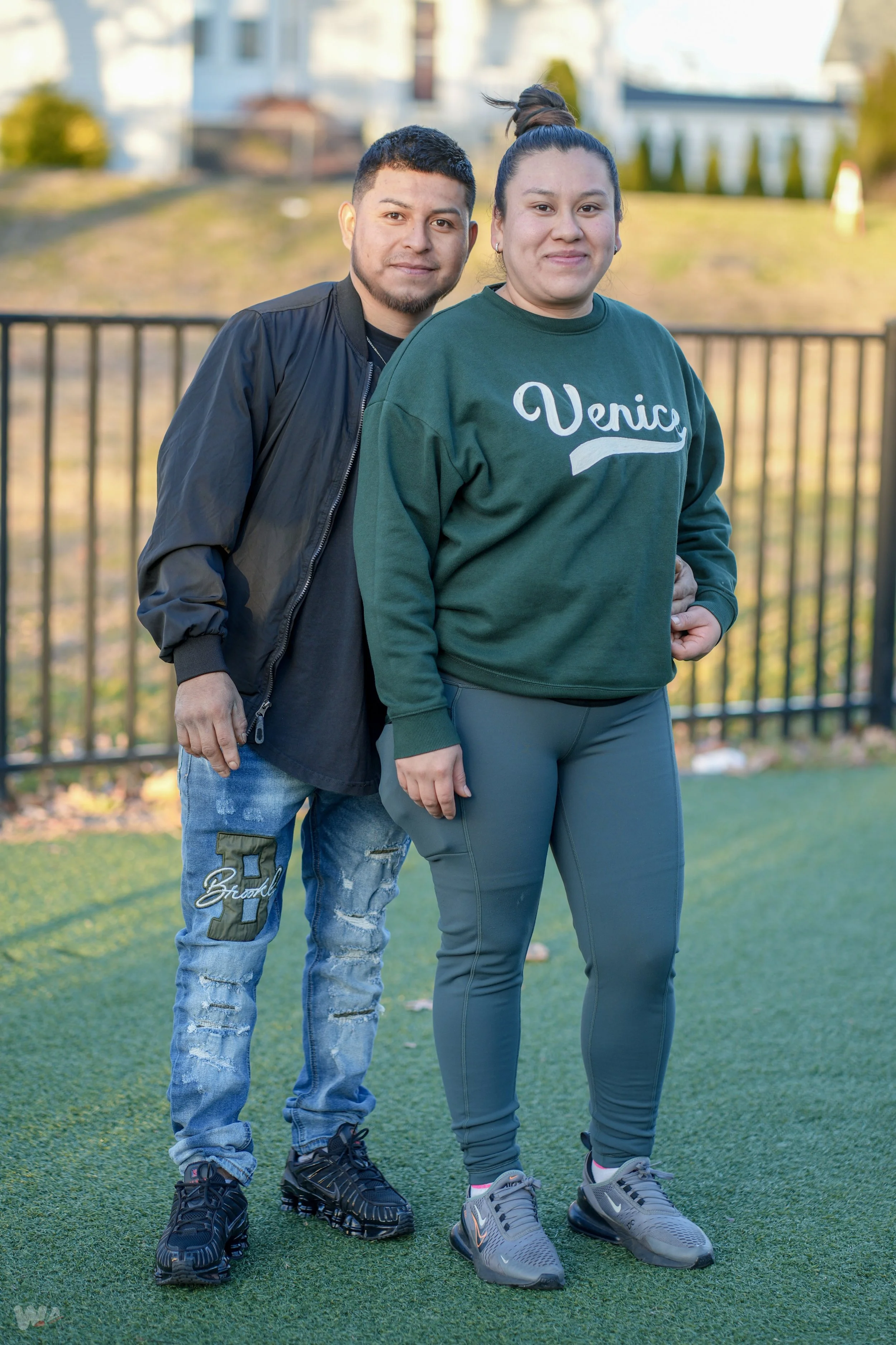 Two people standing outdoors on a field, one man and one woman, both dressed casually and smiling at the camera.