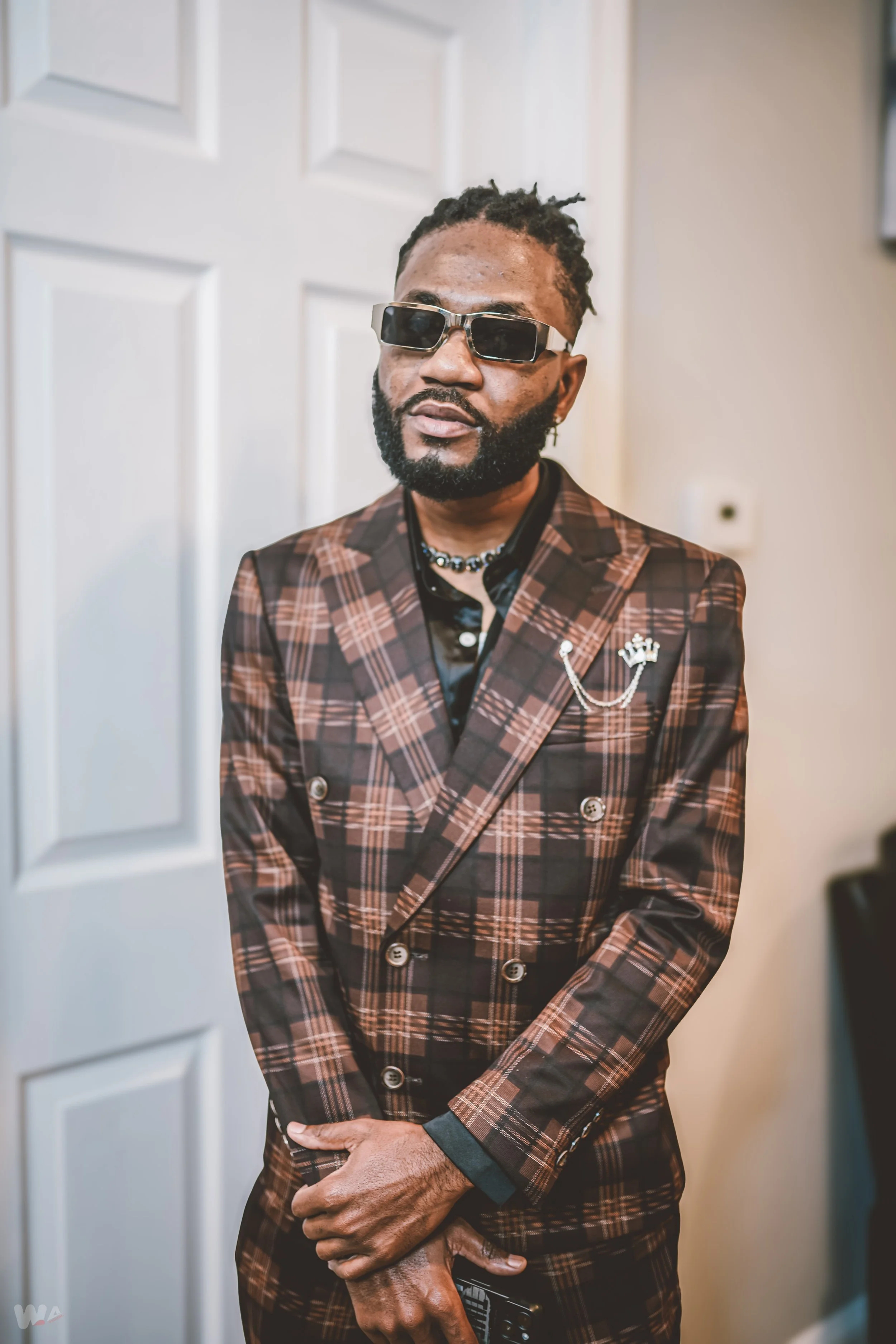 A stylish man with a beard and sunglasses wearing a plaid blazer with a crown pin, black shirt, layered necklaces, standing indoors against a white door.