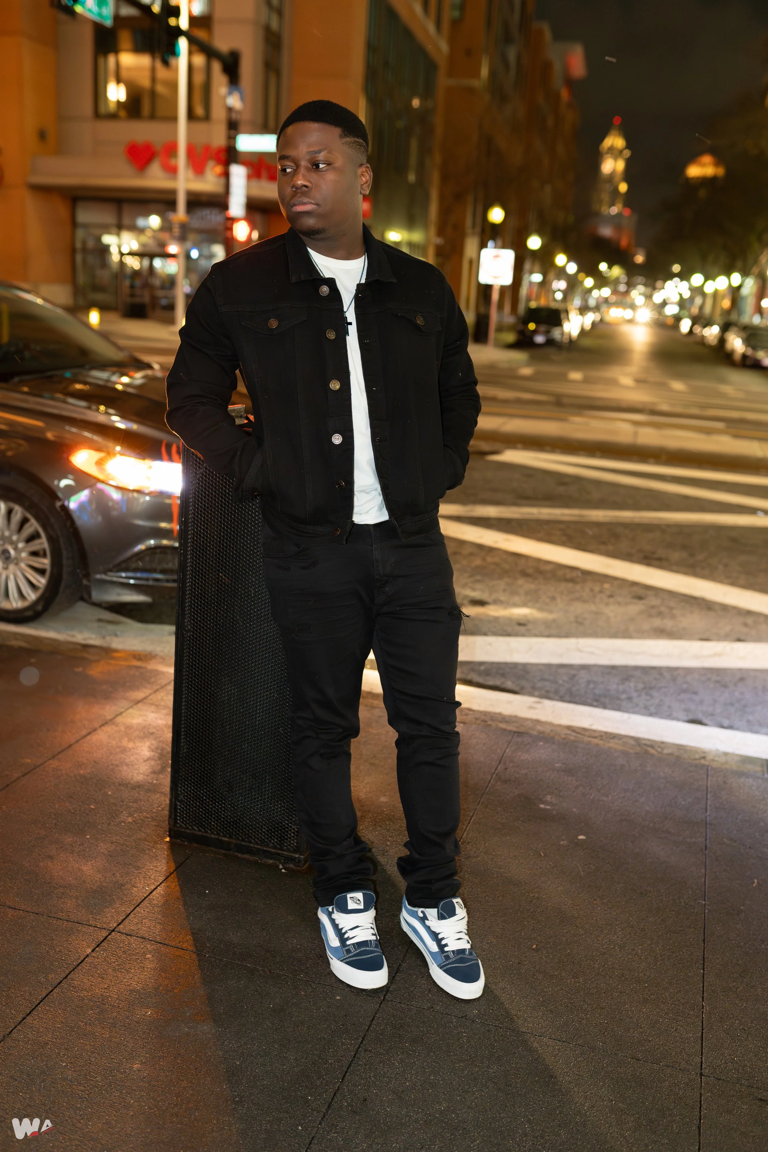A young man in a black denim jacket, black jeans, and sneakers standing on a city sidewalk at night. He is leaning against a black metal barrier with his hands in his pockets, with city lights and buildings in the background.