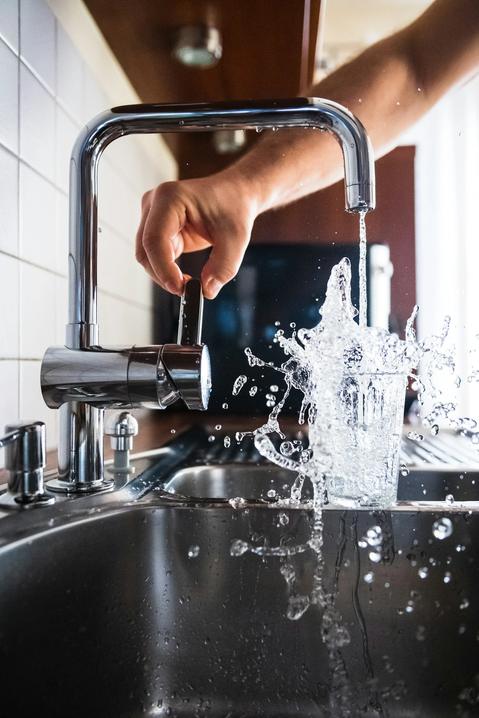 Person turning on a kitchen faucet under running water with a glass catching the water splashing around.