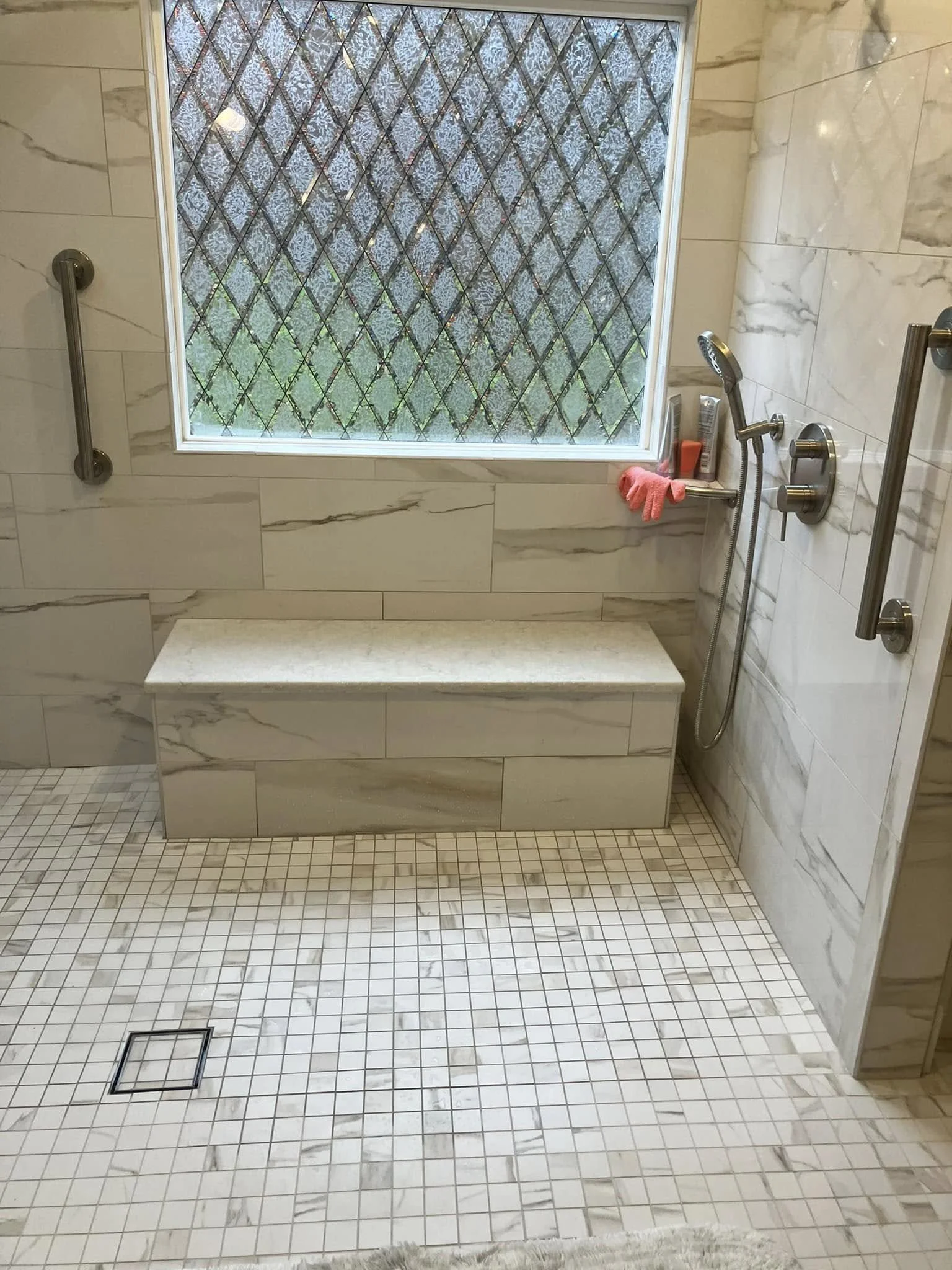 A walk-in shower area with a marble tile bench, white tile walls, small square beige tile floor, a large window with textured glass, and a handheld showerhead with a shelf holding toiletries.