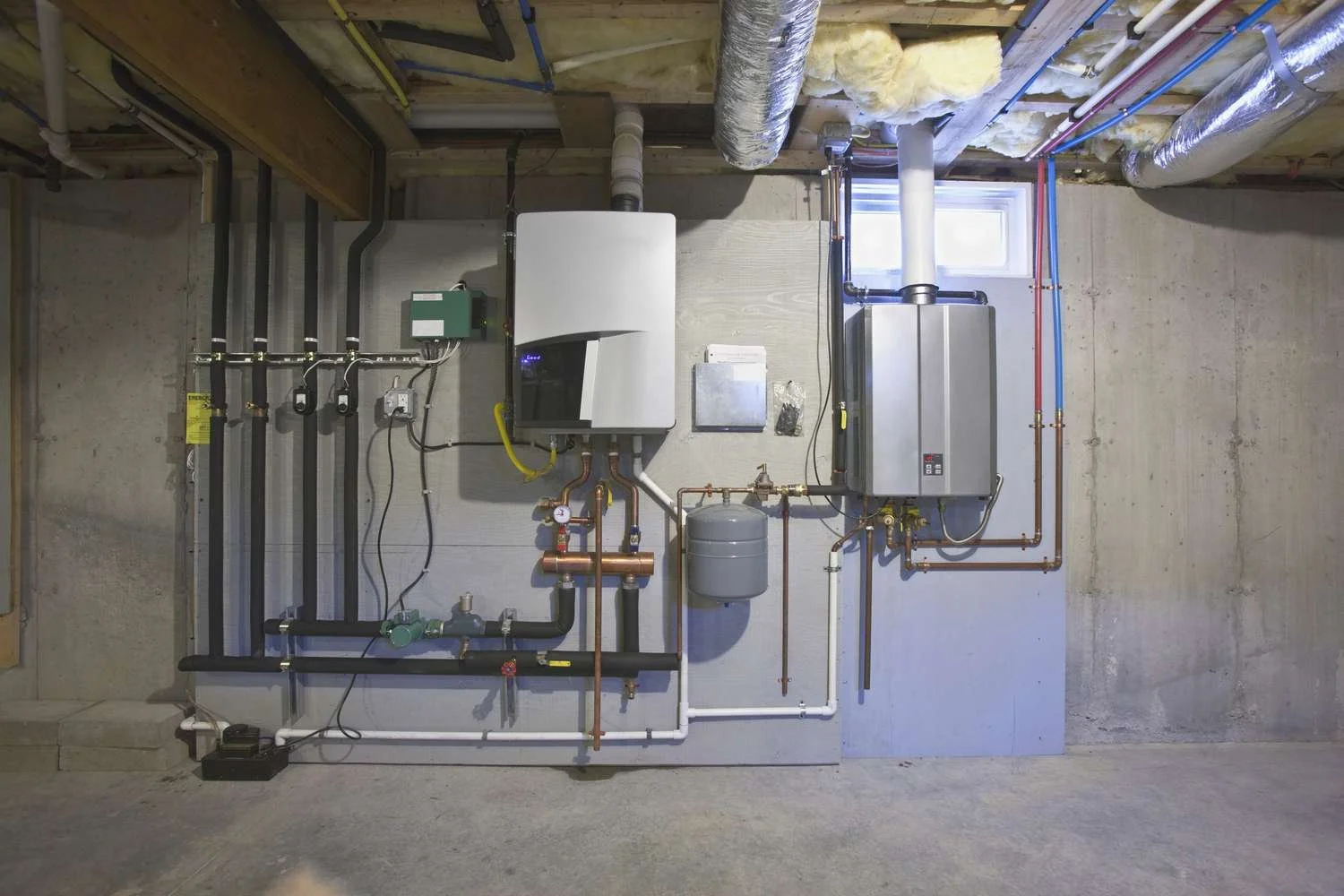 Basement utility room with a water heating and plumbing system including a white tankless water heater, expansion tank, pipes, valves, and electrical connections mounted on a concrete wall.