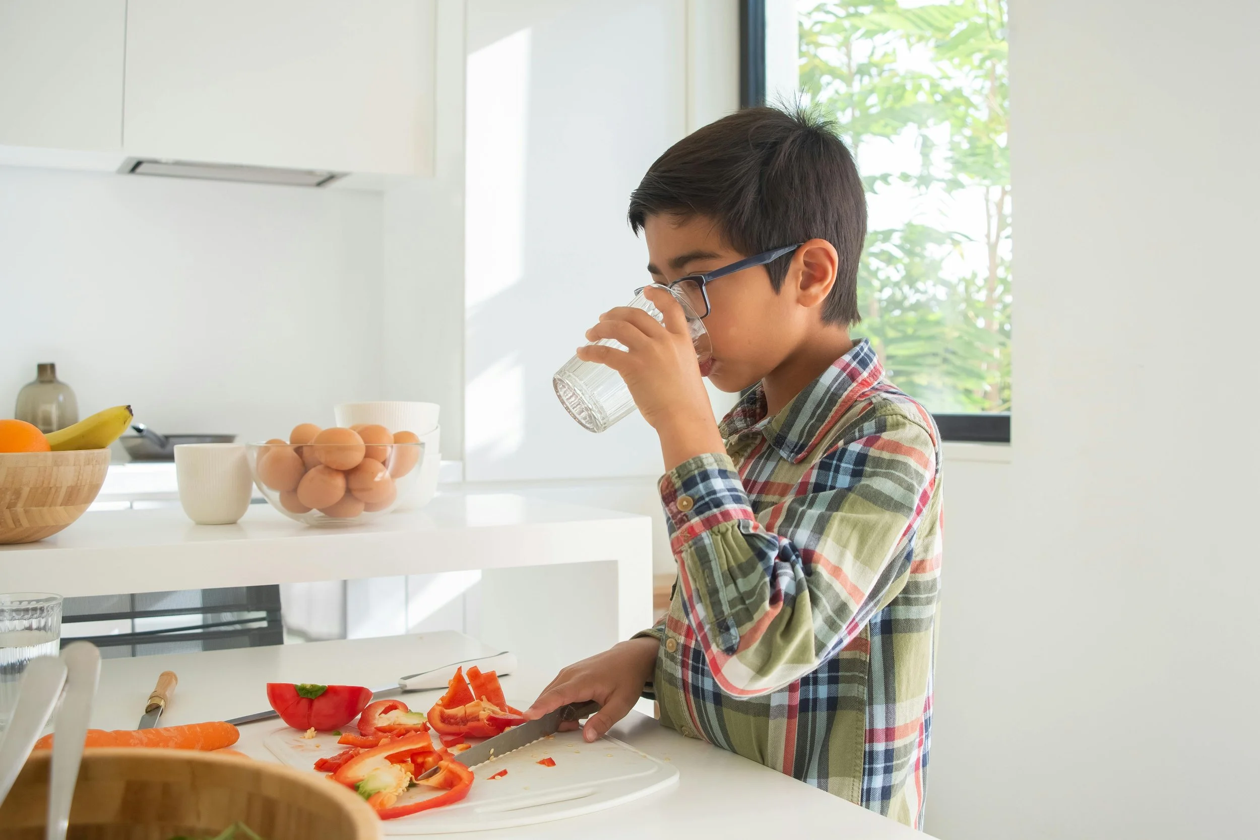 A young boy with glasses, wearing a plaid shirt, is chopping red bell peppers on a white cutting board in a bright kitchen. There are bowls of eggs, bananas, and oranges on the counter, and a window with green foliage outside.