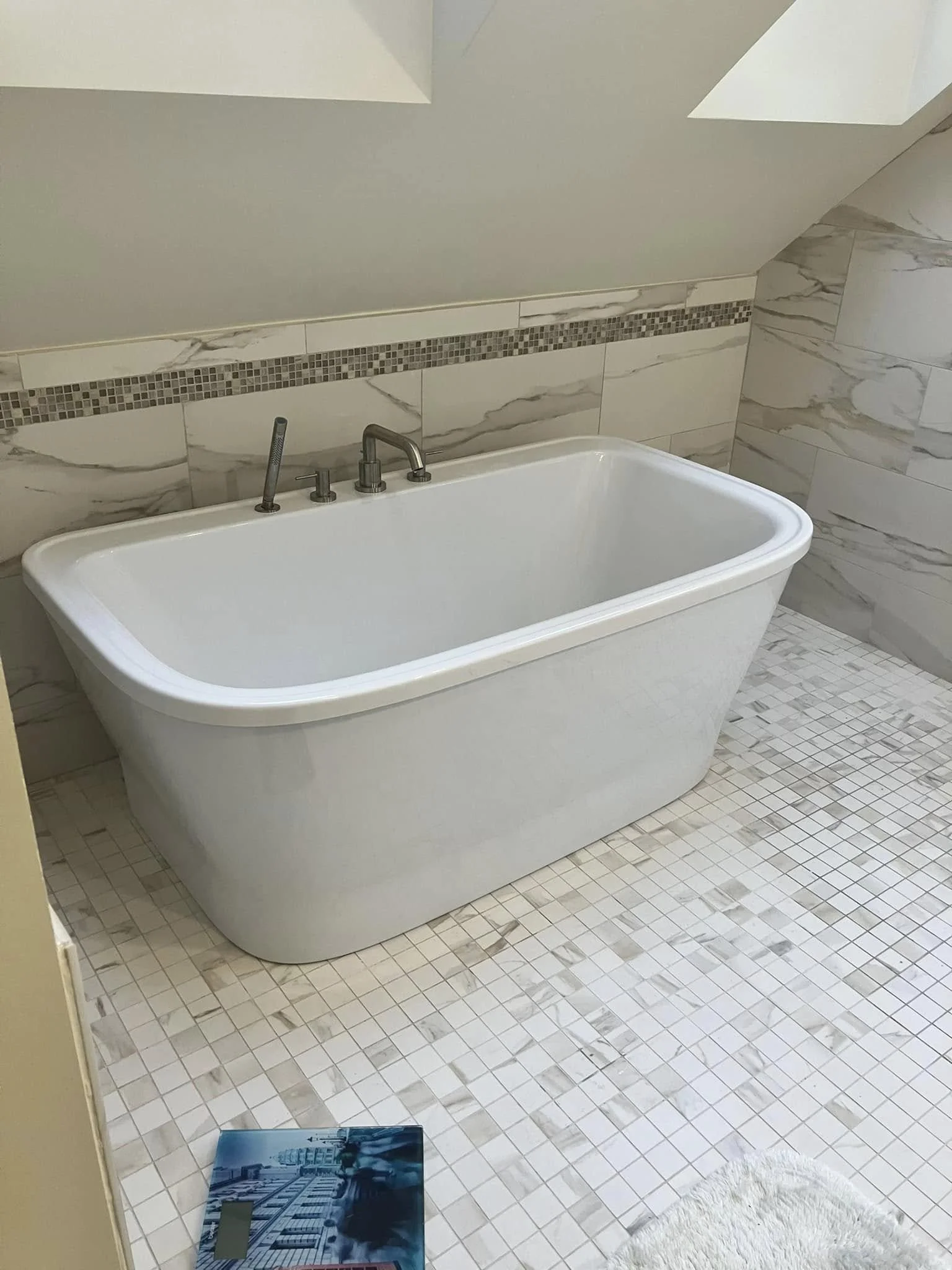 A white bathtub with silver faucet and handles in a bathroom with beige marble tiles and a mosaic border. There is a scale and a white rug on the tiled floor.