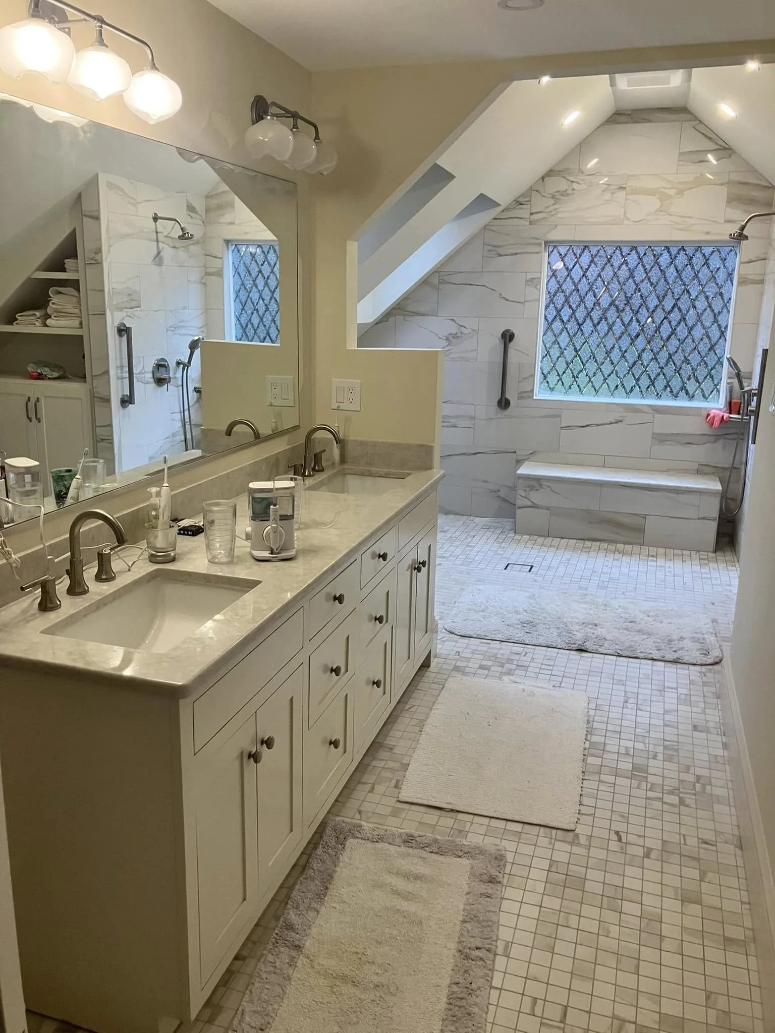 A spacious bathroom with a double sink vanity, large mirror, and a walk-in shower with a bench and window, decorated in neutral tones and tile flooring.